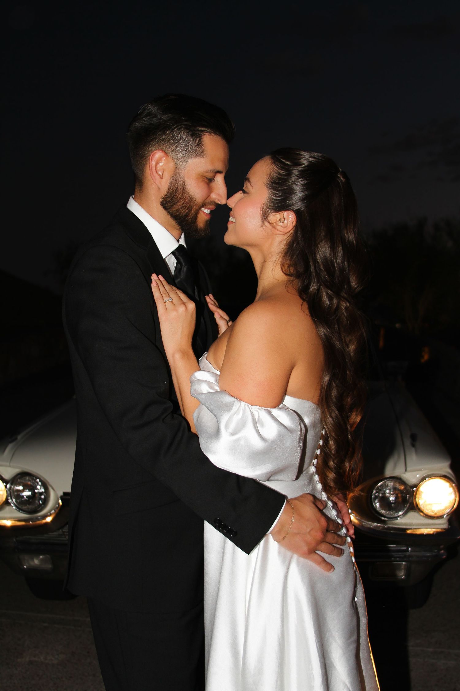 Romantic nighttime portrait of couple in formal attire near vintage headlights.