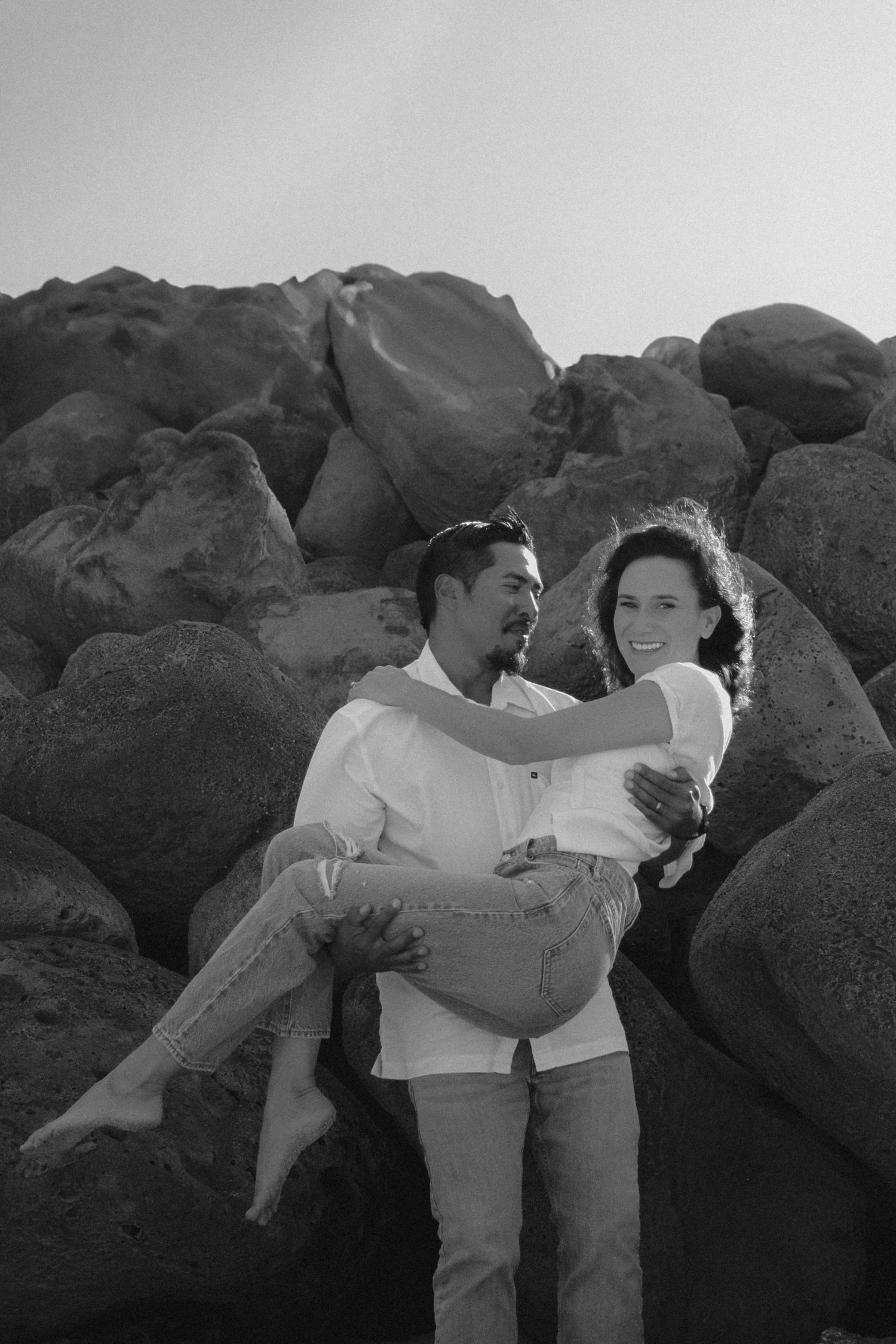 Tyler Garlej Photography - A romantic couple sits together on rocky coastal boulders in a black and white photograph.