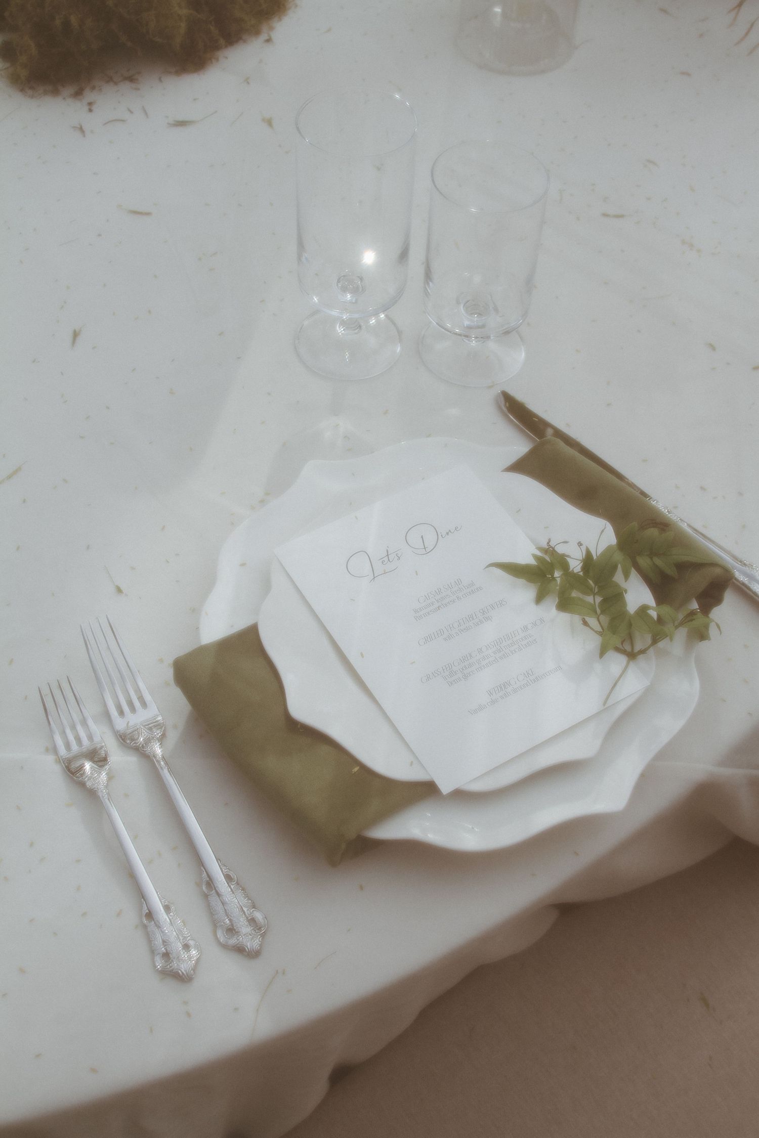Elegant wedding place setting with olive green napkin, menu card, and fresh greenery on white linen.