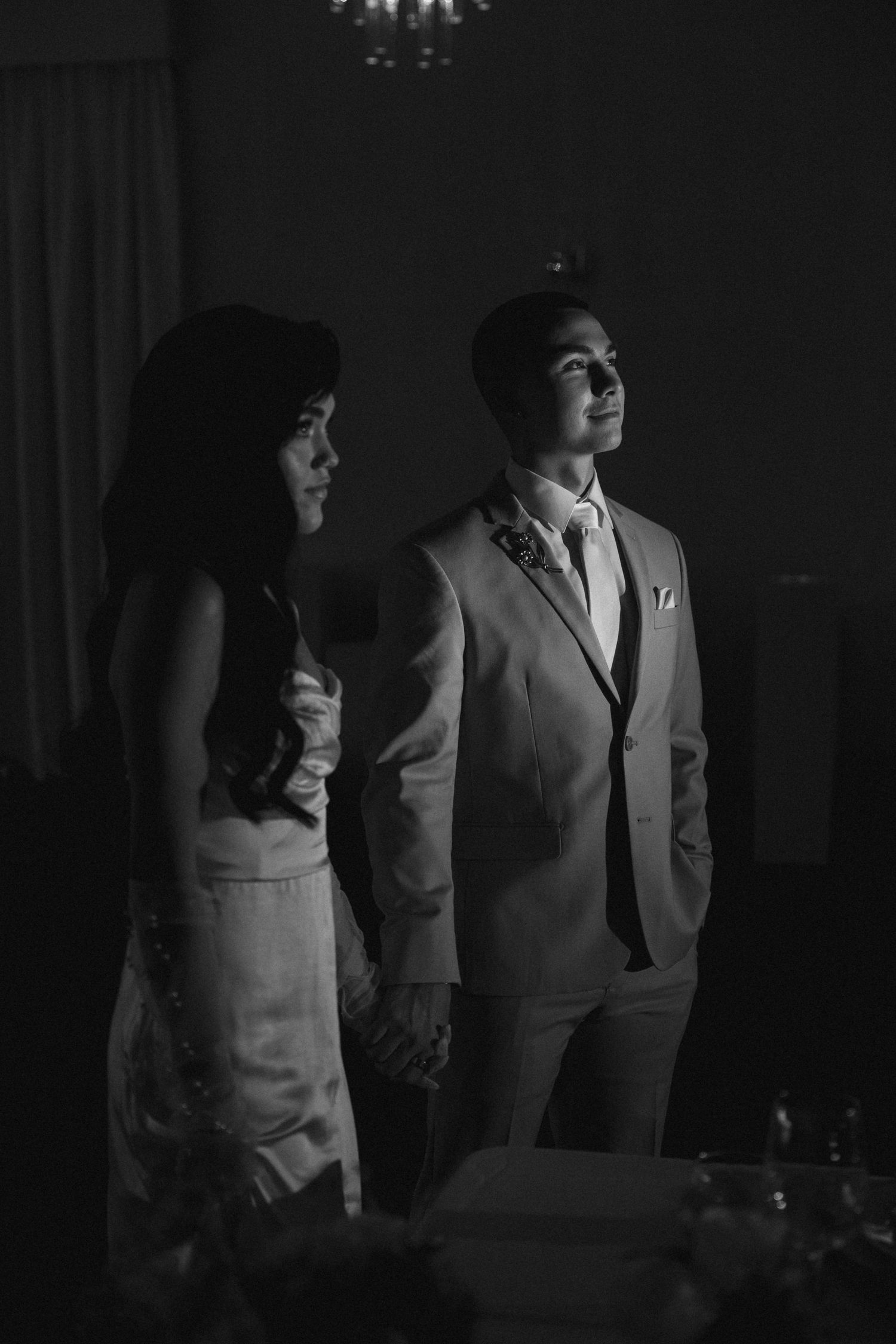 Black and white photo of an elegant couple holding hands at a formal wedding reception in a dimly lit venue.
