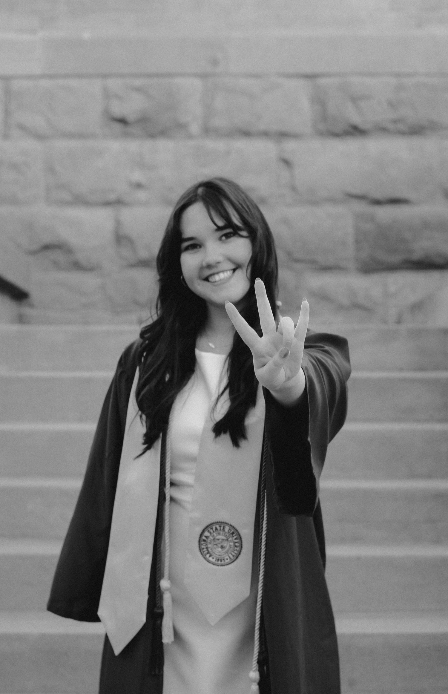 Graduate making peace sign while wearing graduation cap and gown on stone steps.