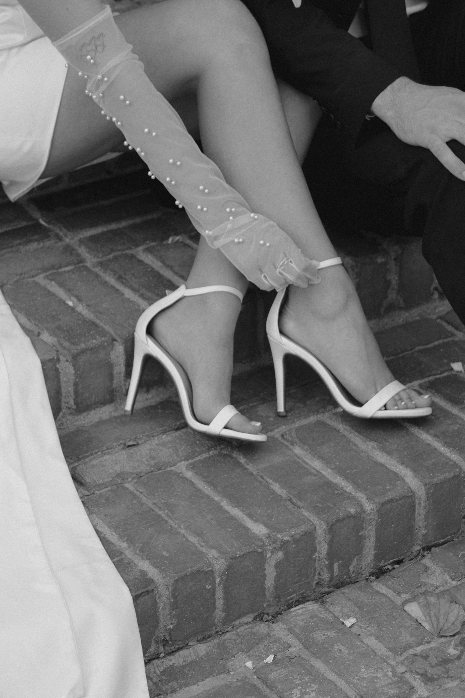 Black and white photo of a bride wearing white strappy heels and pearl-embellished gloves sitting on brick steps.