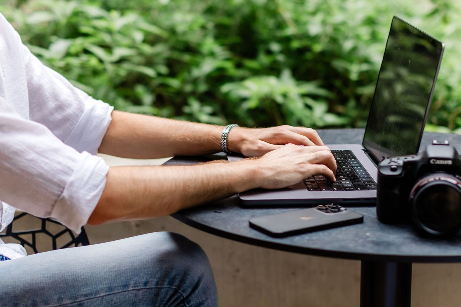 Person working outdoors on laptop with camera on table in leafy green garden setting.