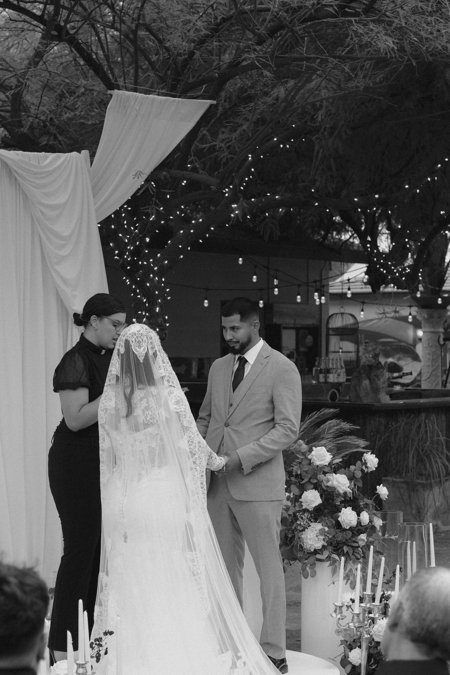 A black and white wedding ceremony photo under a draped arch with twinkling lights in the background.