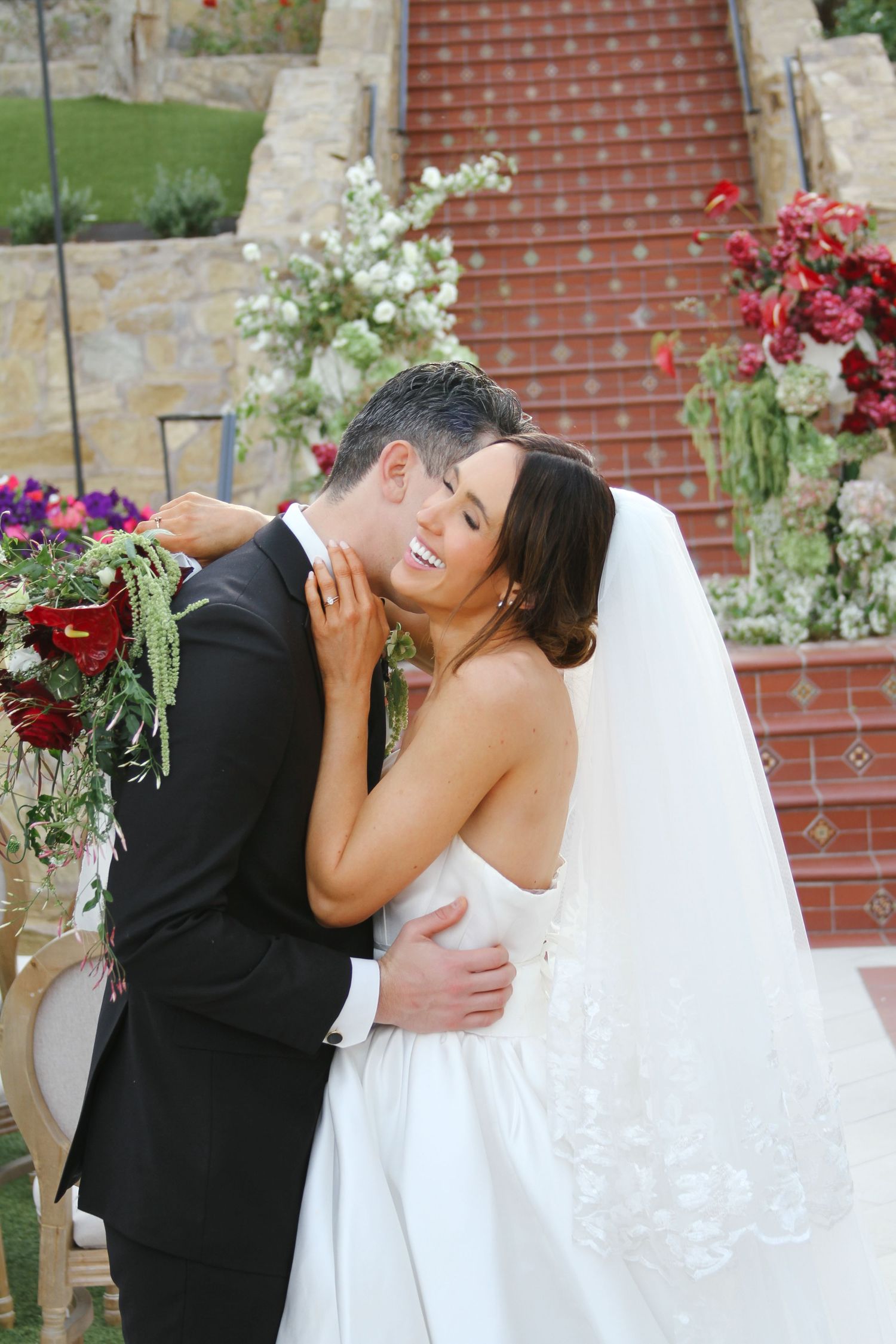 A bride and groom share a romantic embrace outdoors, with floral arrangements and stone steps in the background.