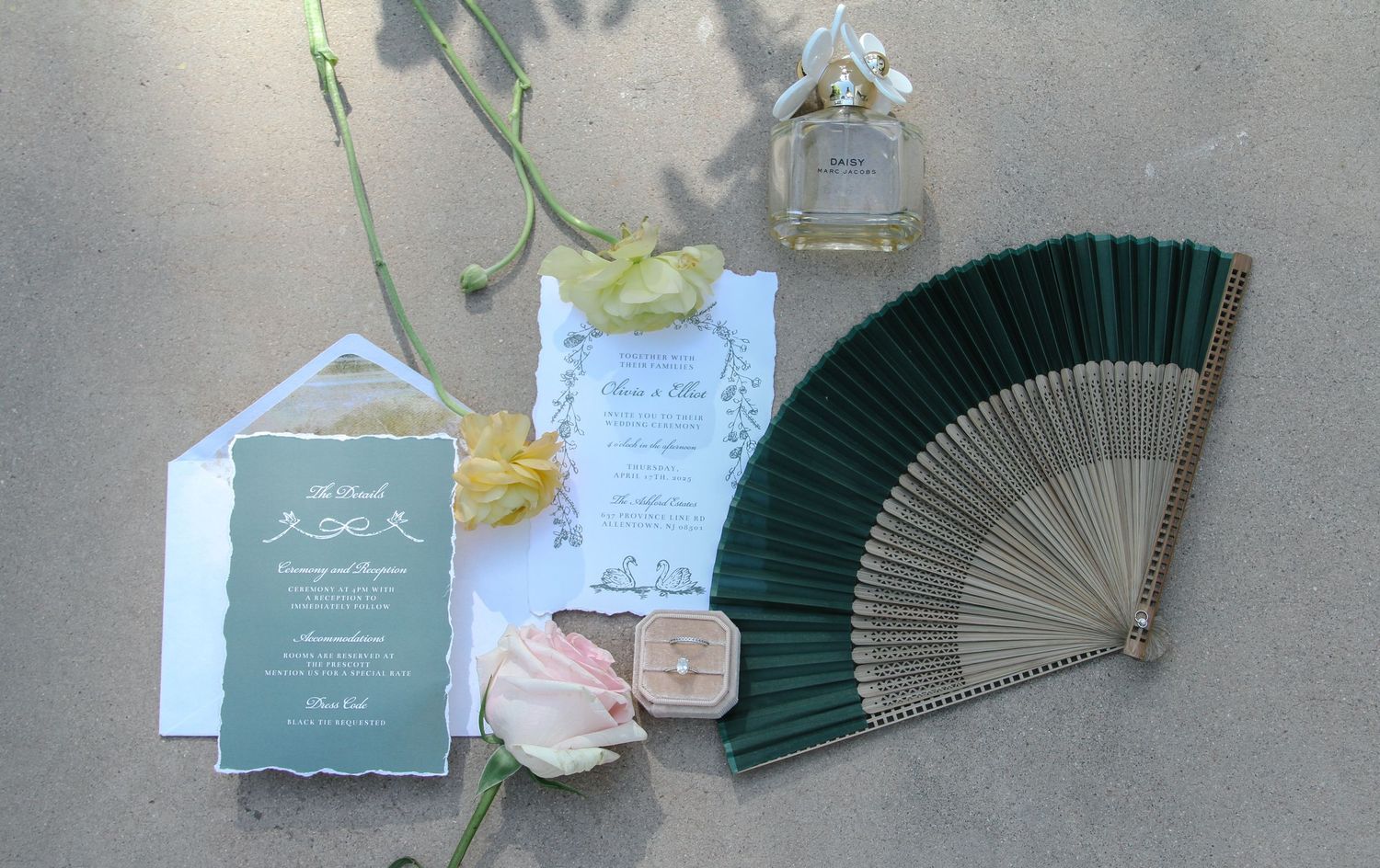 Wedding invitation suite with a dark green folding fan, pink roses, and a perfume bottle arranged on a gray surface. Tyler Garlej Photograph