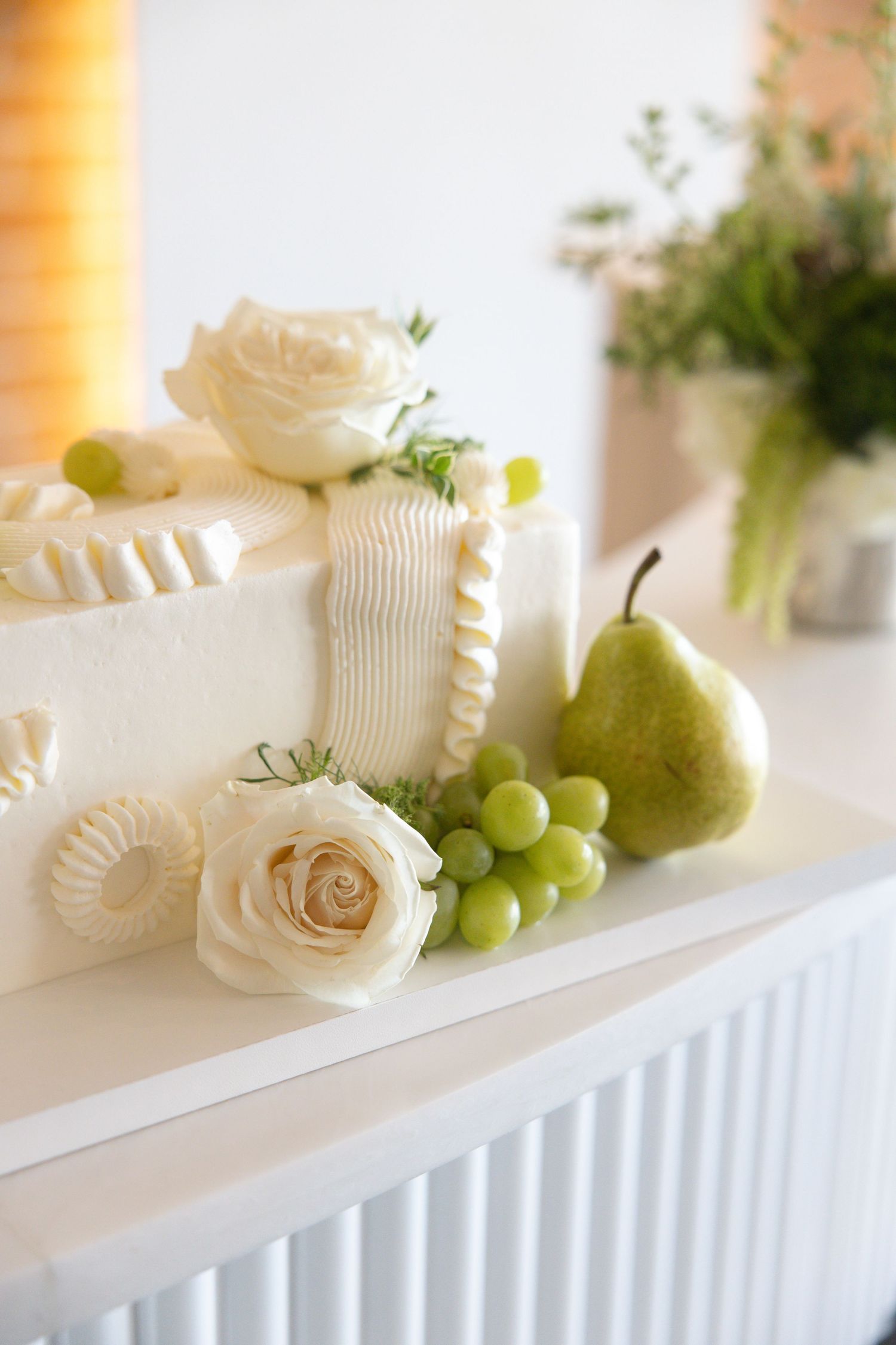 Elegant white wedding cake decorated with cream roses, green grapes, and a fresh pear on display.