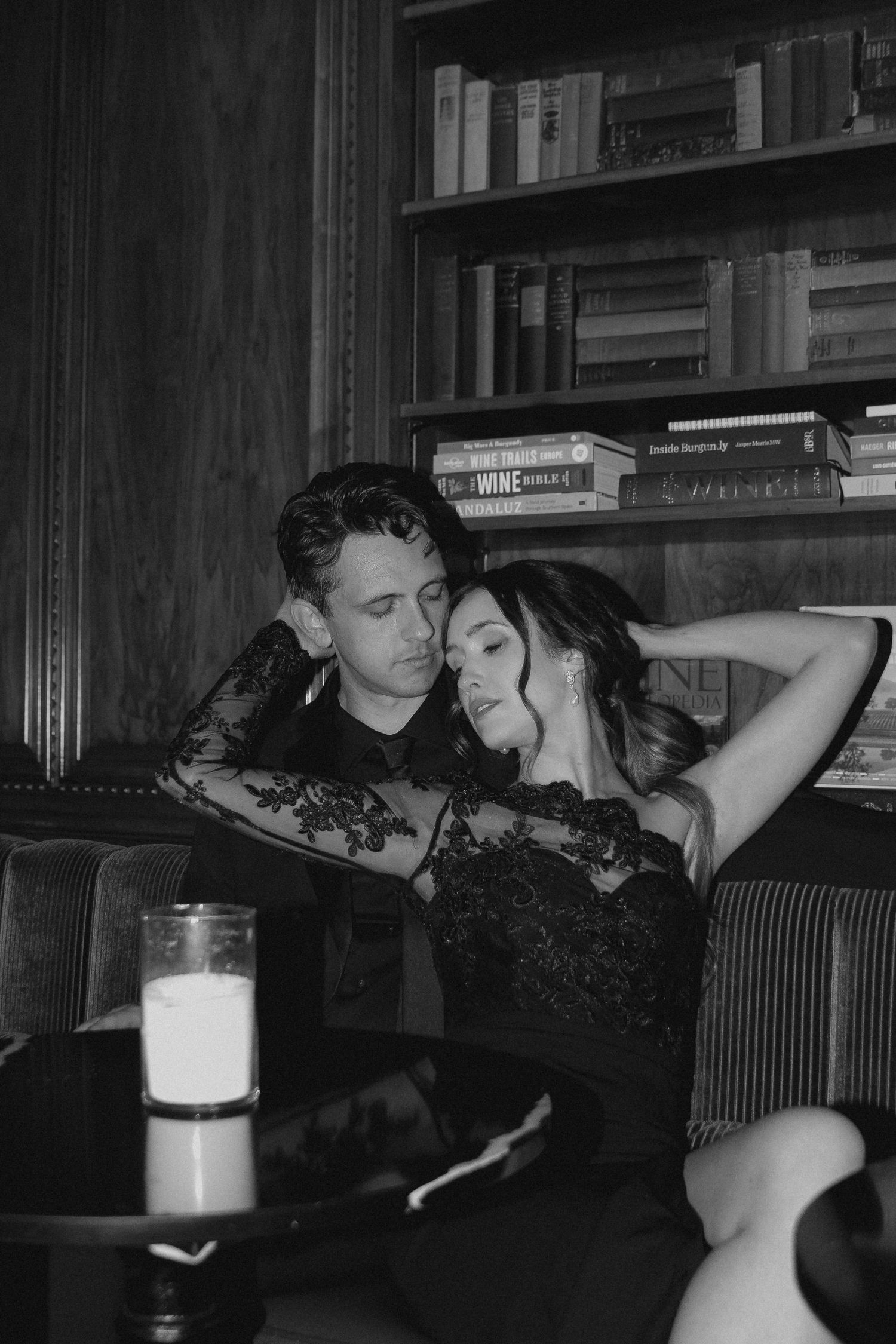 A romantic black and white photo of a couple in lace embracing in front of a bookshelf at a dimly lit venue.
