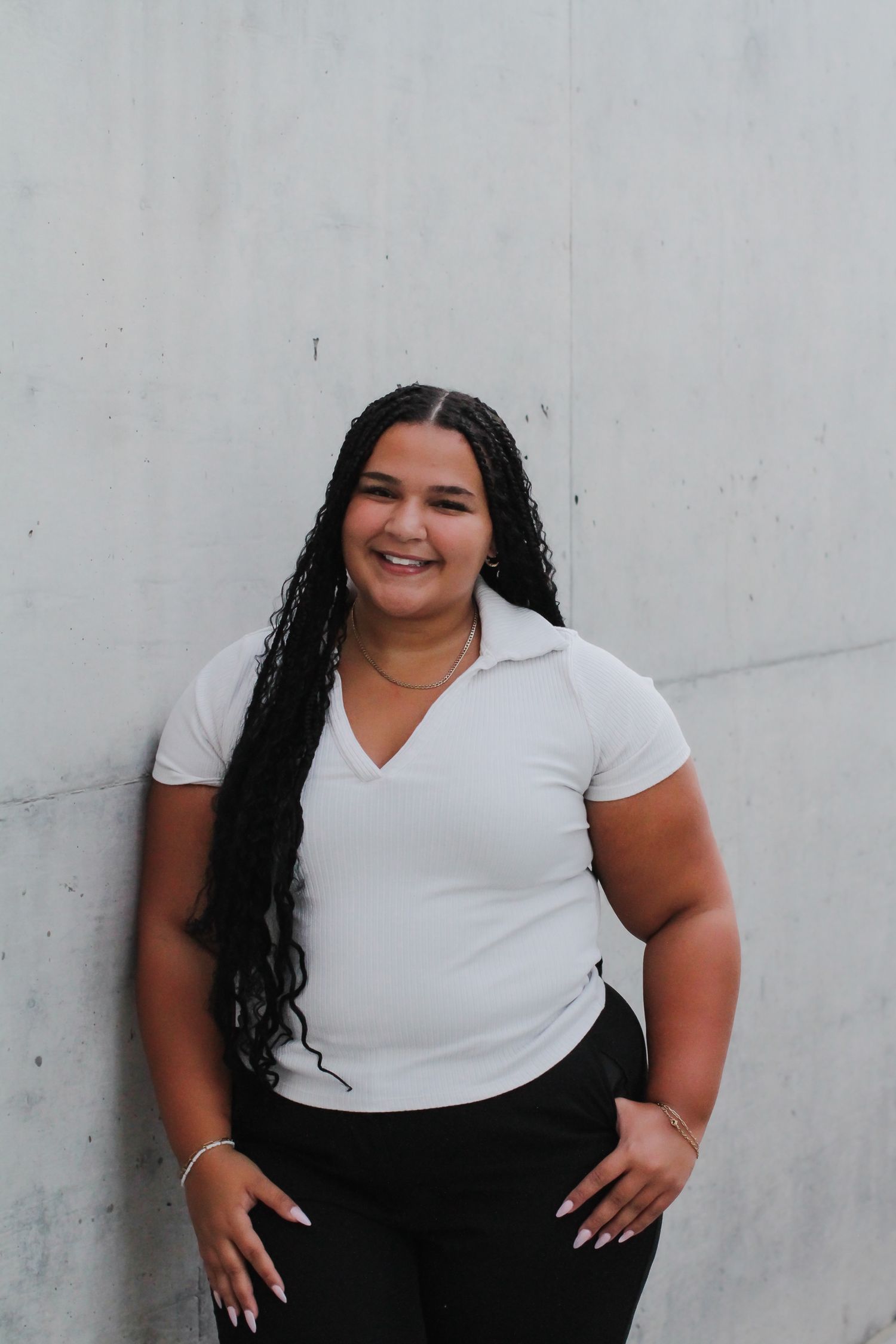 A person with long dark hair wearing a white shirt and black pants poses against a grey wall.
