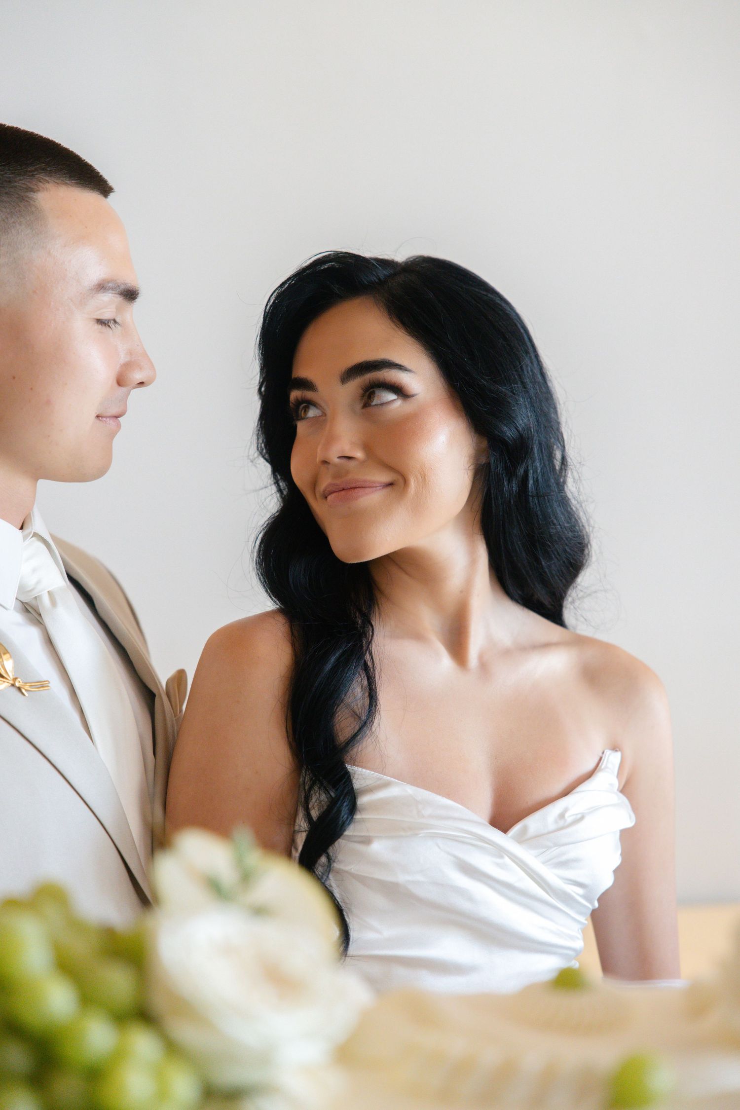 Bride in white strapless gown gazes lovingly at groom in tan suit at elegant wedding reception table.