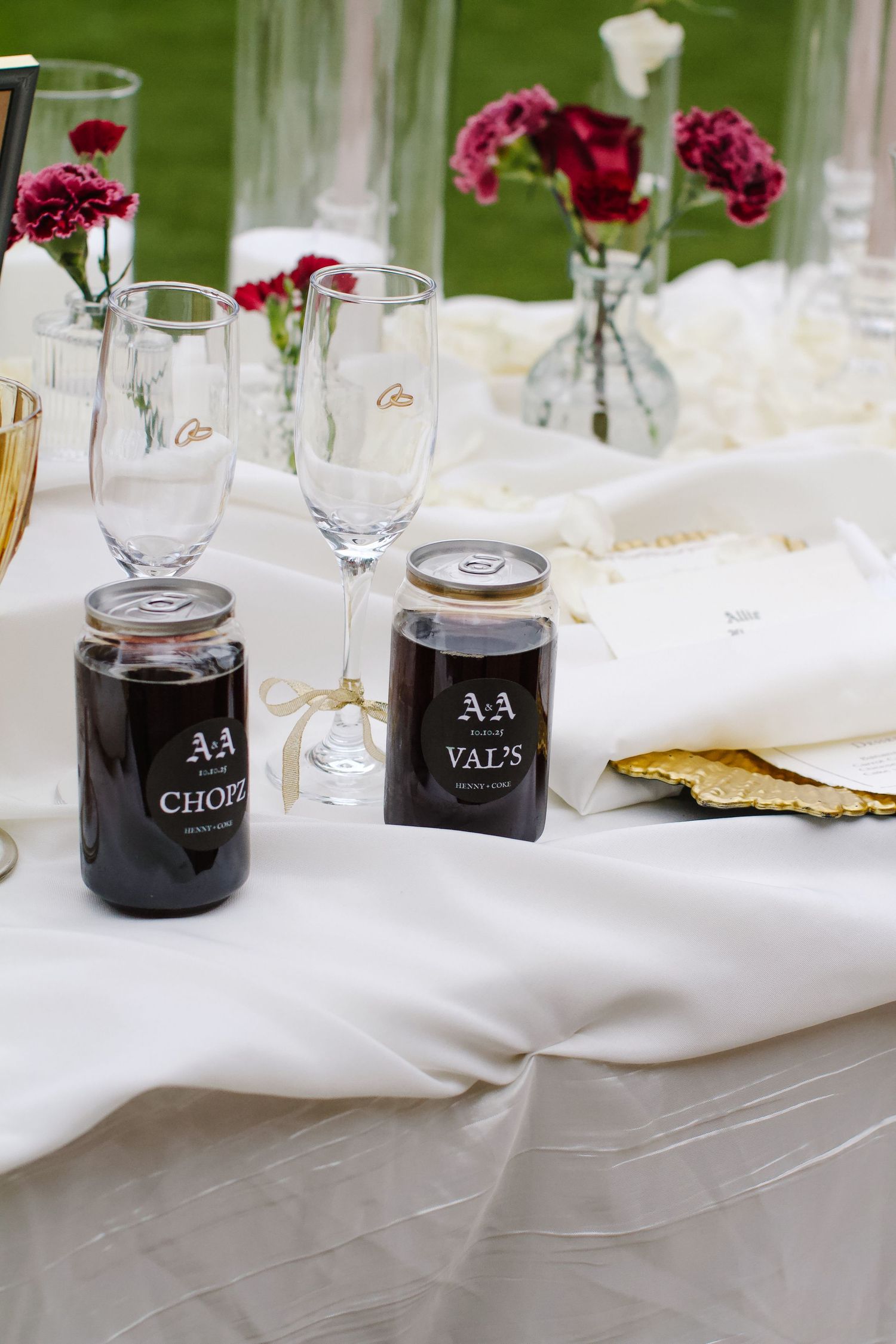 Close-up of wedding table settings with labeled black jars, wine glasses, and burgundy floral accents on white linens.