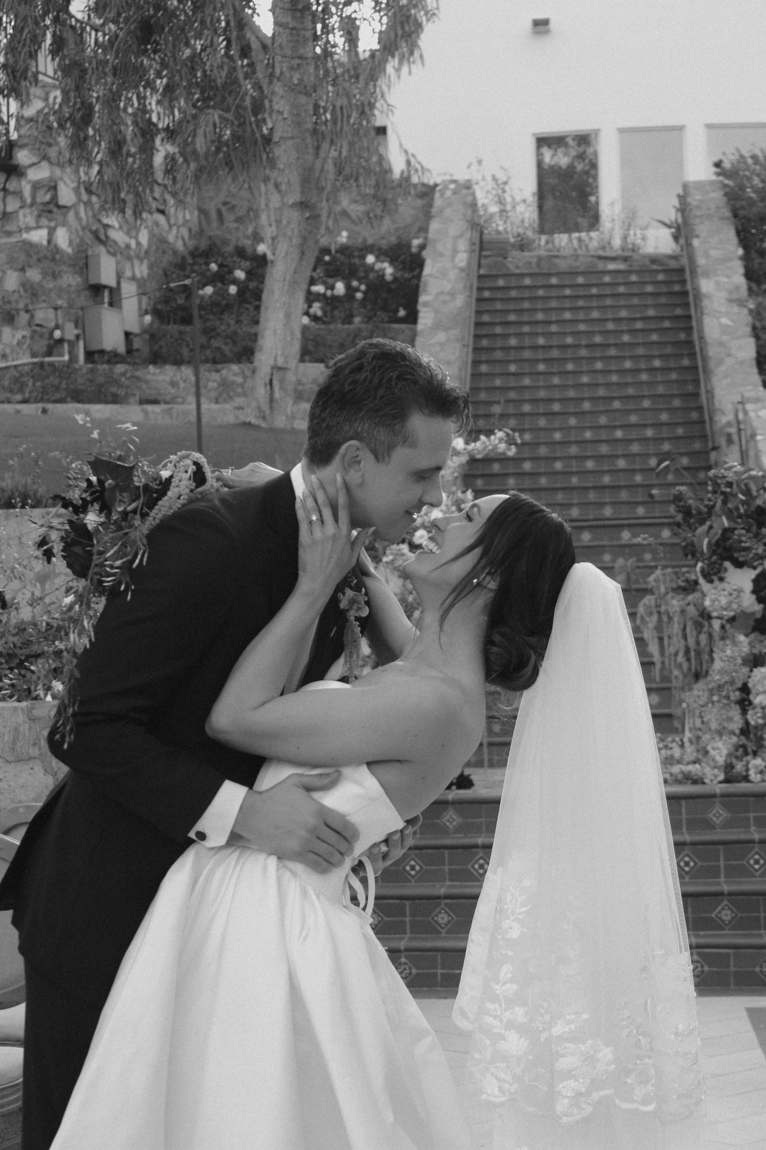 Black and white photo of a bride and groom sharing a romantic kiss outdoors near decorative stairs on their wedding day.