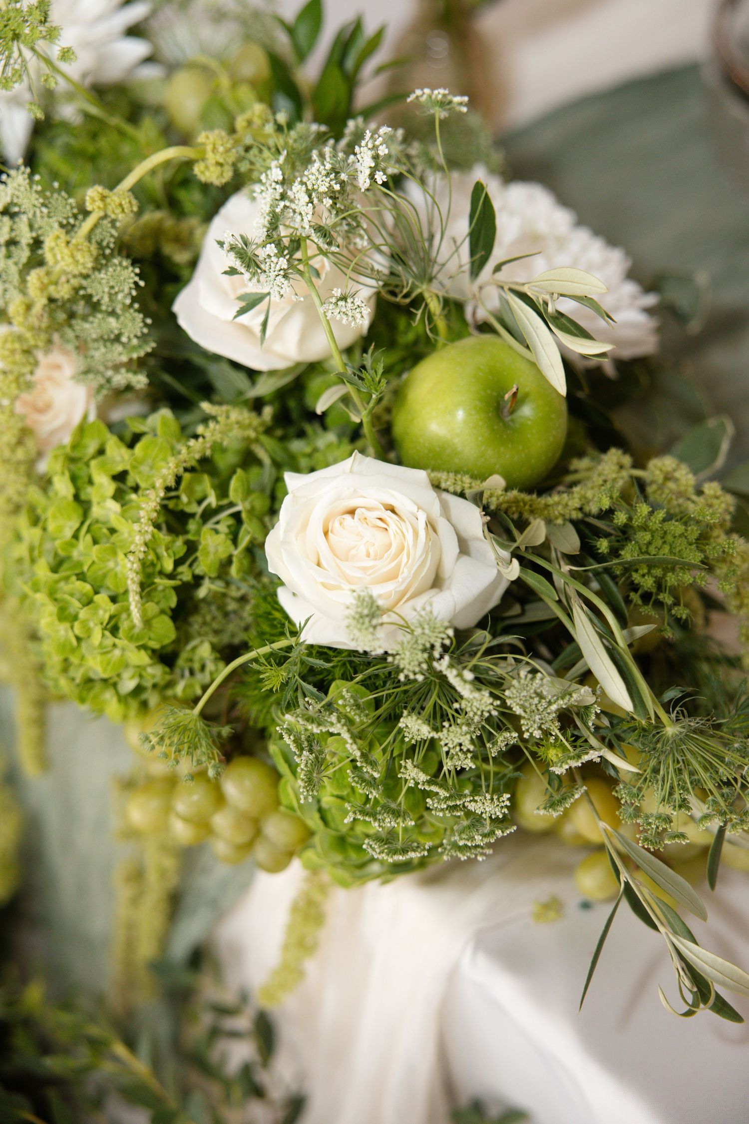Elegant floral arrangement featuring white roses, green apples, grapes, and lush greenery for a wedding centerpiece.