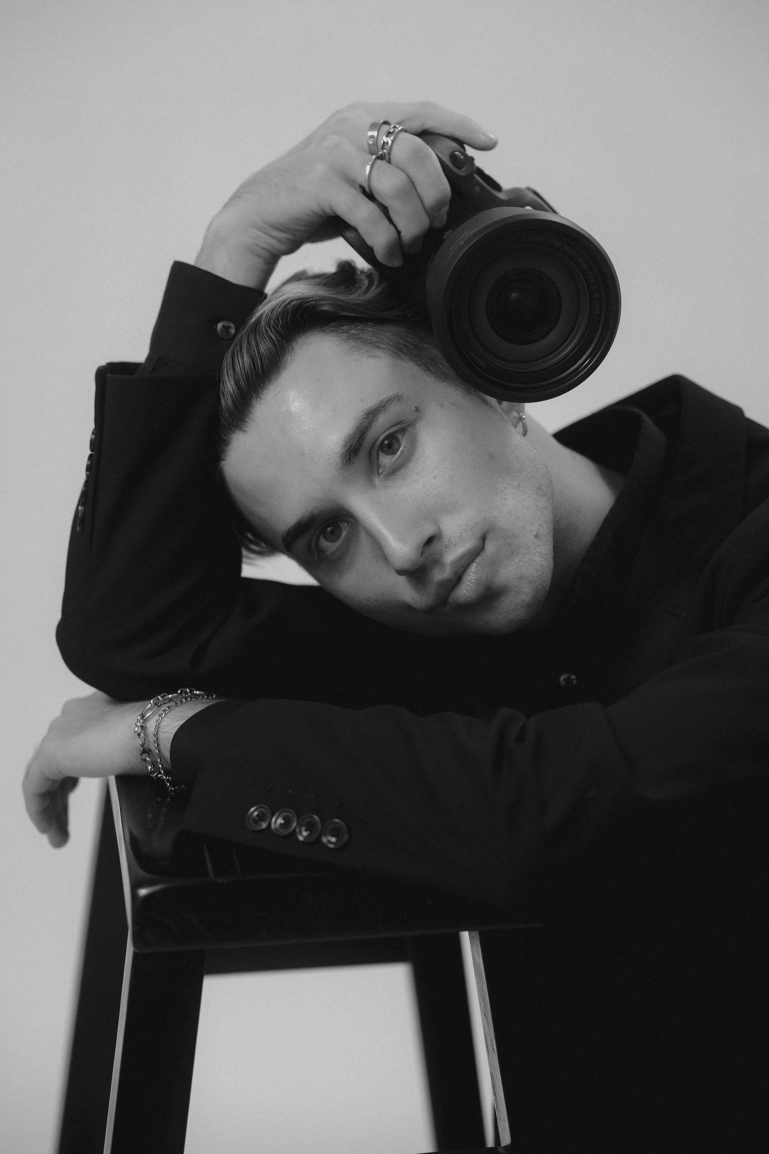 Black and white portrait of a young man in black jacket holding a camera on his head, leaning on a stool.