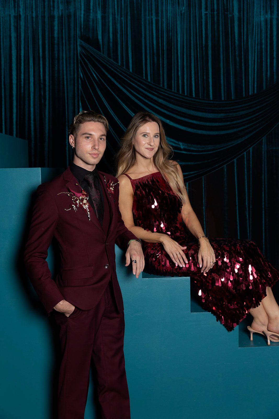 A young couple poses formally, man in burgundy suit, woman in red sequin dress, against teal velvet backdrop.