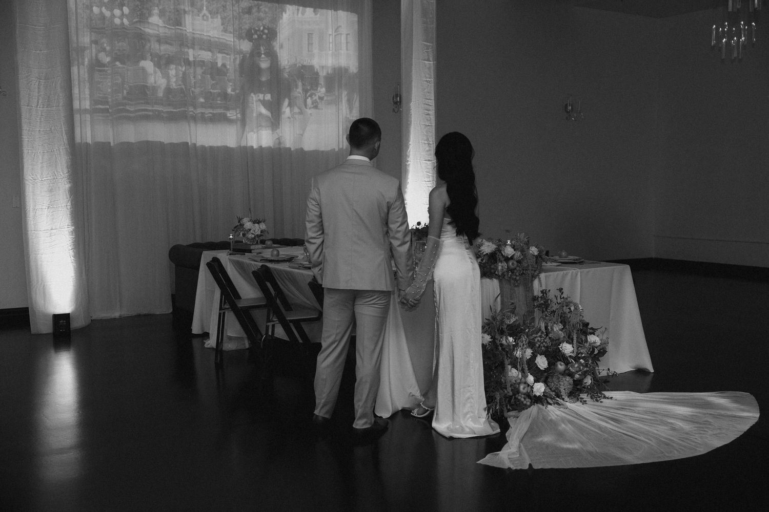 Black and white photo of bride and groom holding hands, viewing wedding reception decor from behind.
