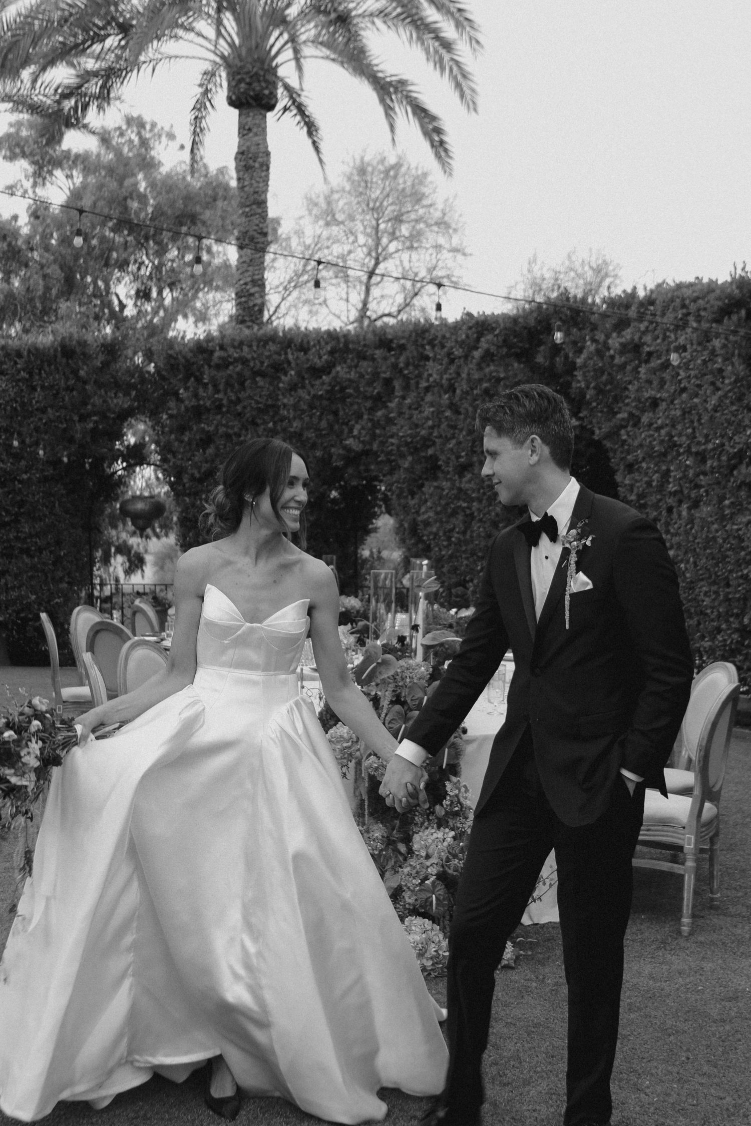 Black and white photo of smiling bride in strapless gown and groom in tuxedo holding hands outdoors.