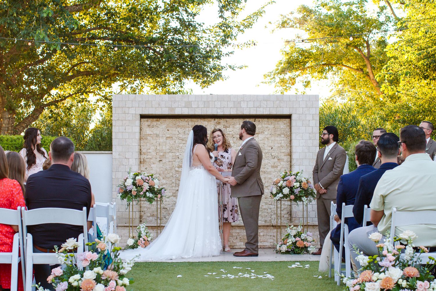 Wedding ceremony guests gather outdoors on a sunny day with white floral arrangements and chairs set up on a lawn.