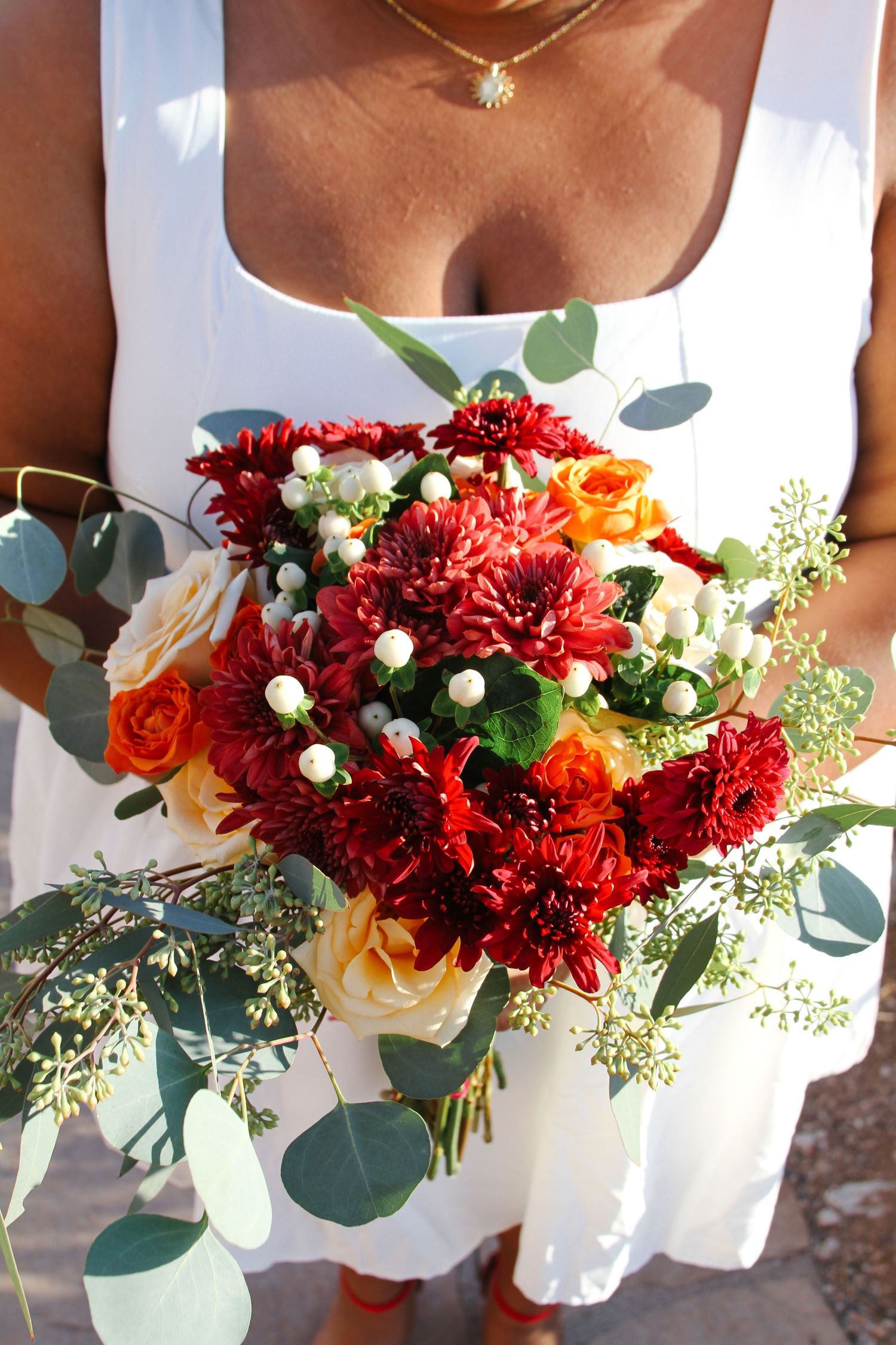 Beautiful red and orange bridal bouquet with eucalyptus leaves against white dress.