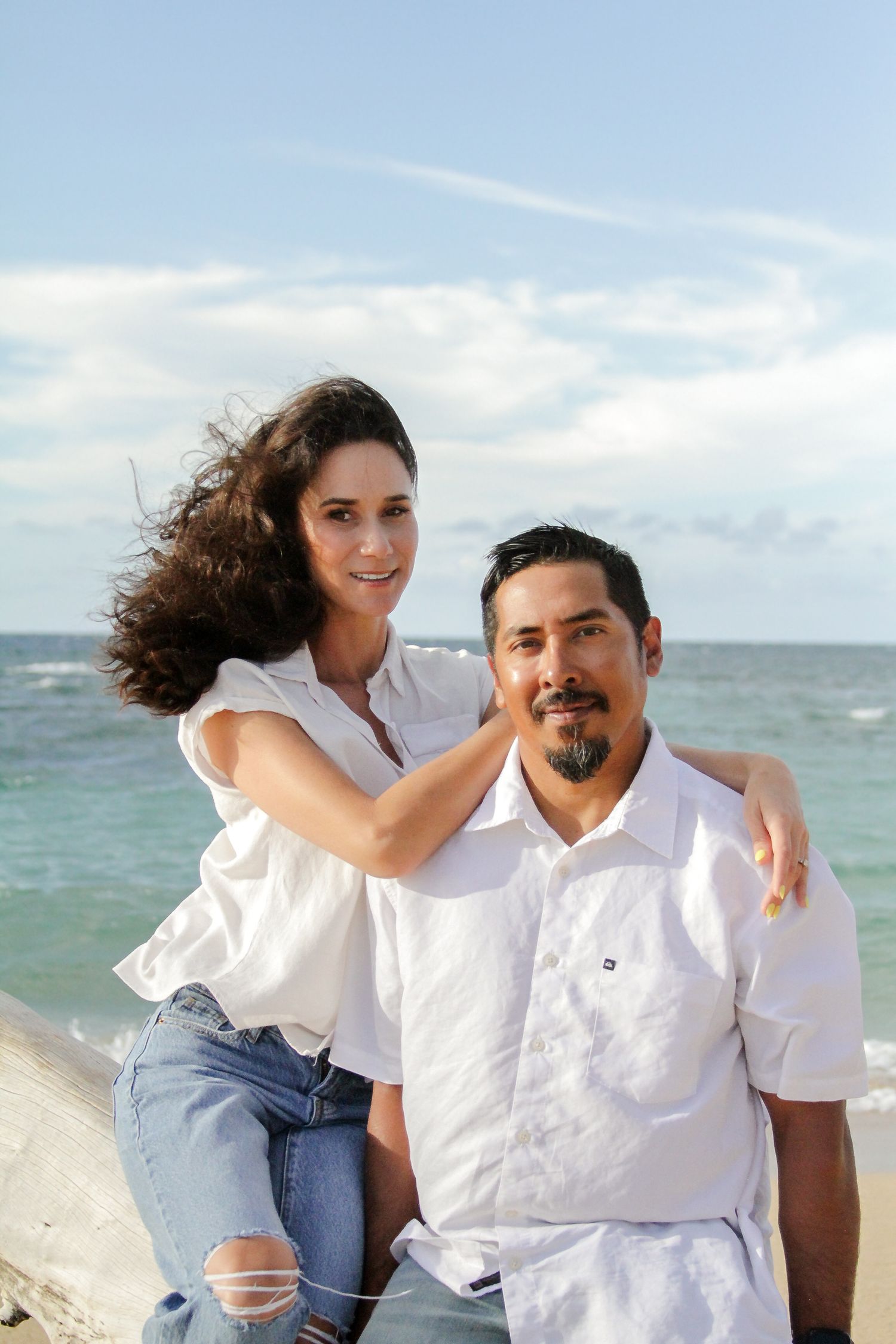 A couple in white shirts poses together at the beach with ocean waves and blue sky in the background.