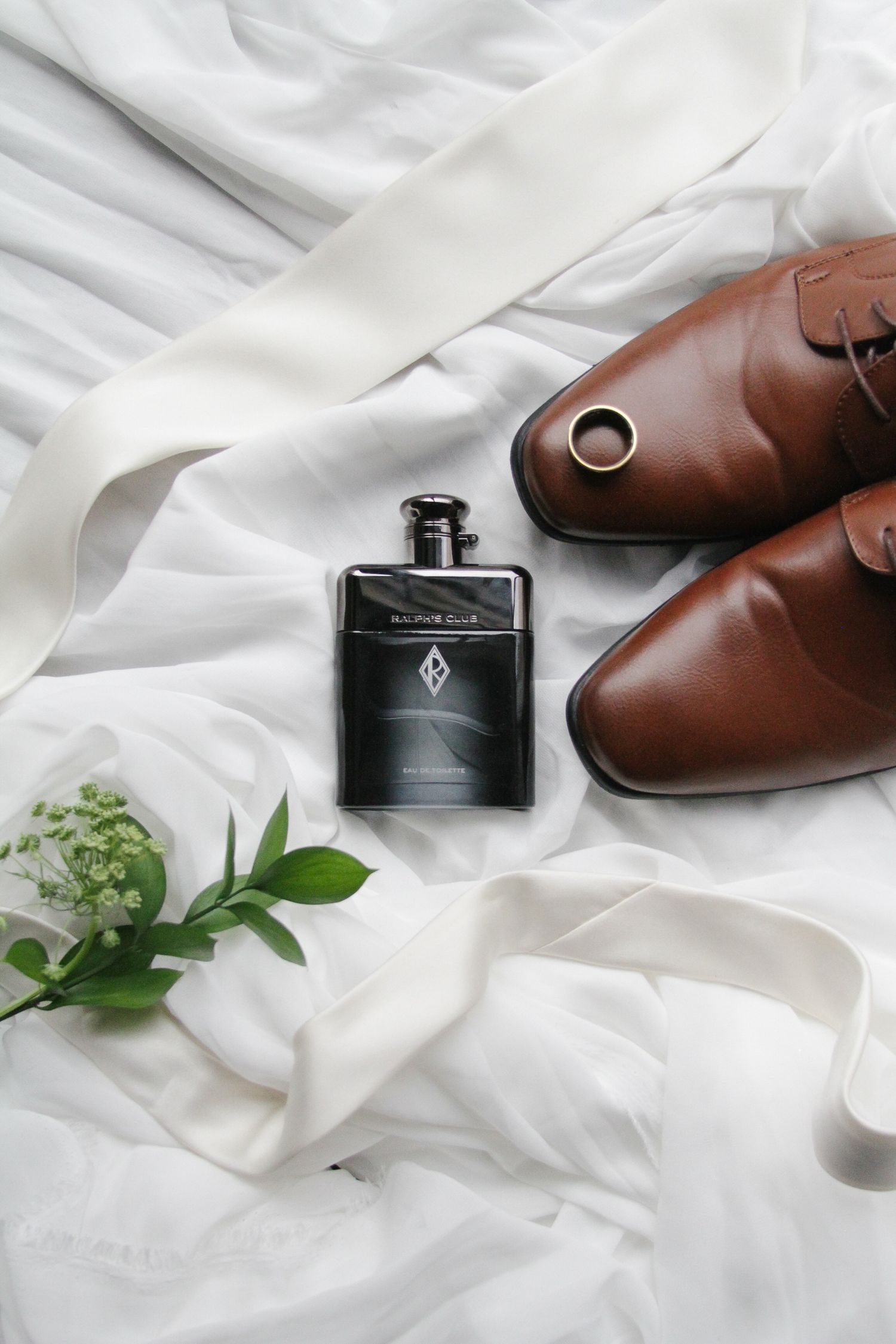 Wedding flat lay with brown leather monk shoes, black Ralph Lauren cologne, gold ring, and white floral sprig on white fabric.