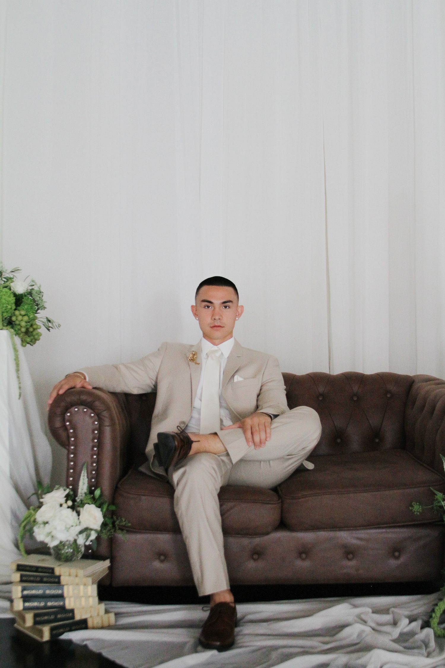A person in a beige suit sits casually on a brown tufted leather couch against a light wall with decorative plants.