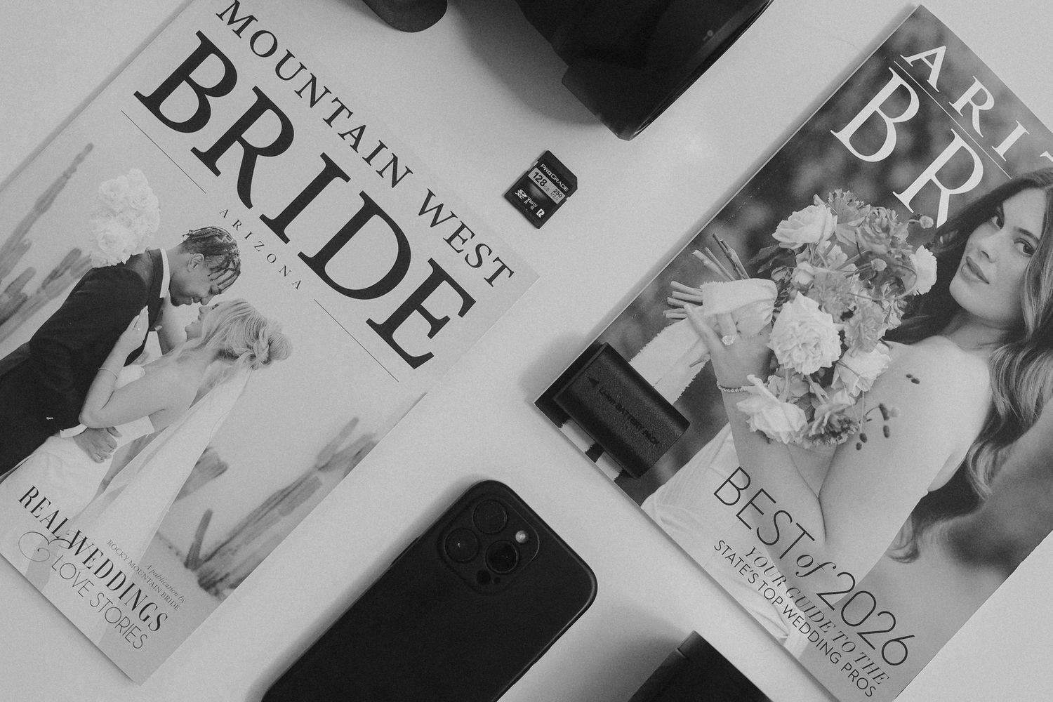 Black and white photo of Mountain West Bride and Art Bride magazines with a camera and smartphone on a white surface.