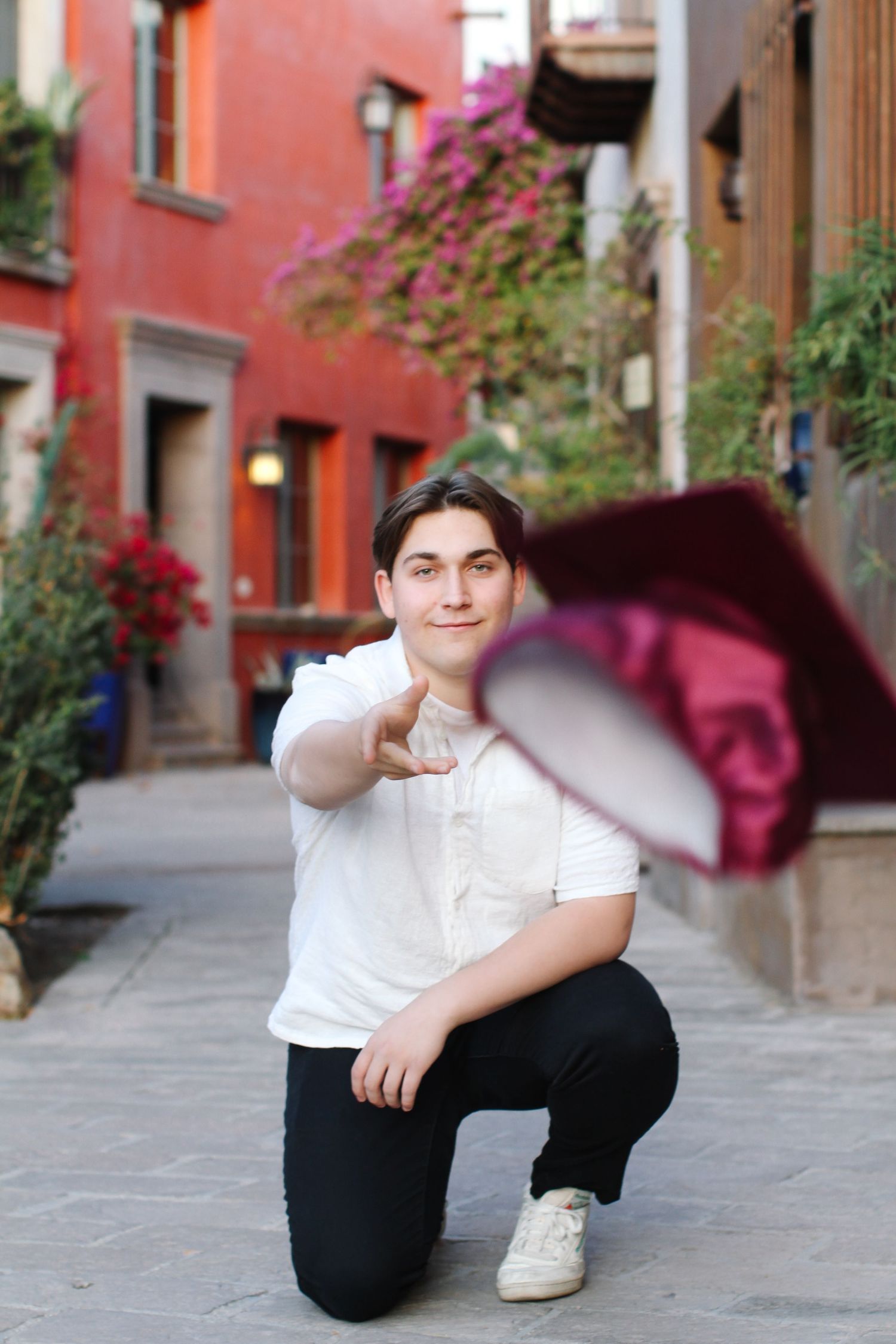Graduate in white shirt kneeling on a cobblestone street with colorful buildings and flowers.