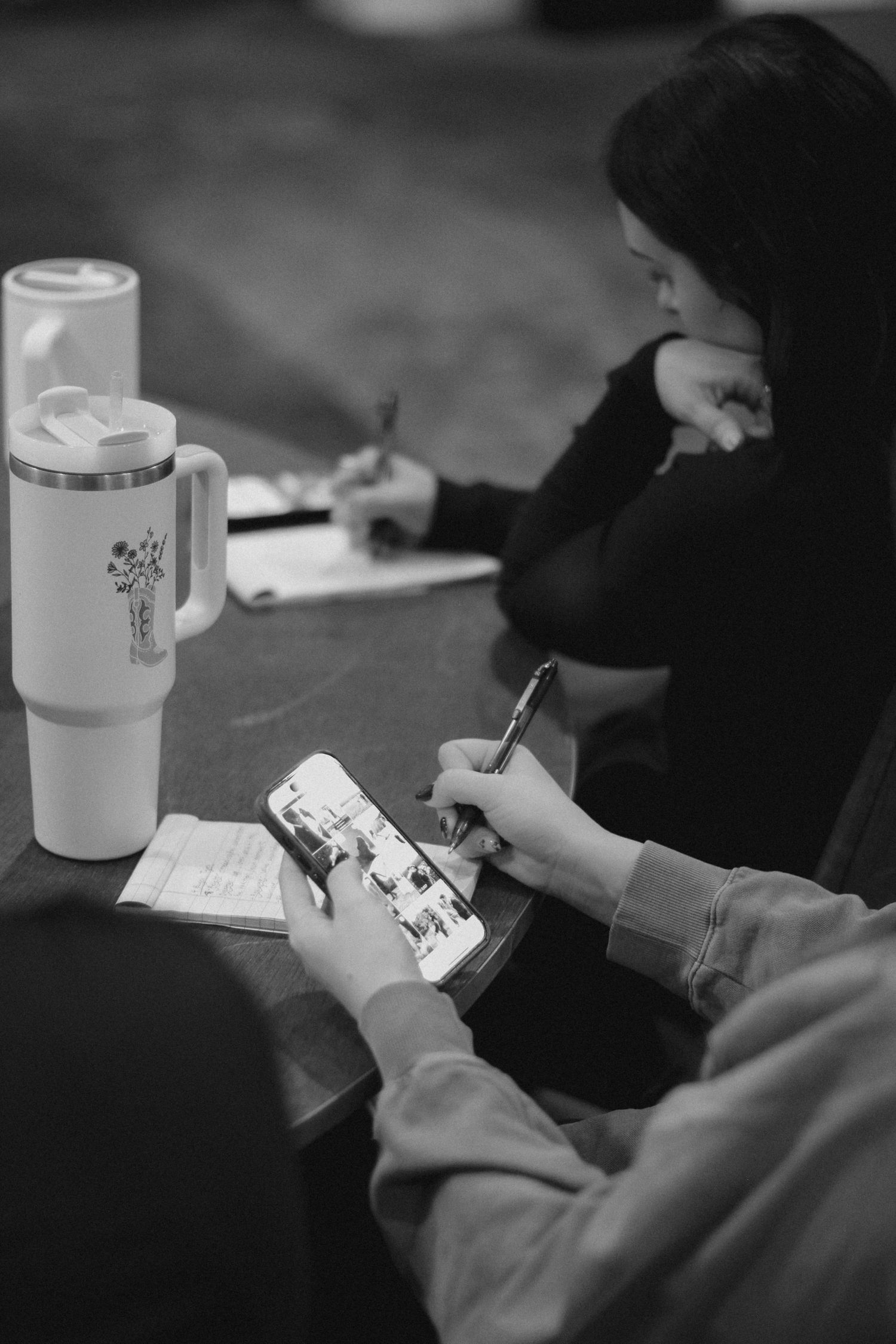 Black and white photo of two women at a table, one using a phone, one writing, with a Stanley tumbler nearby.