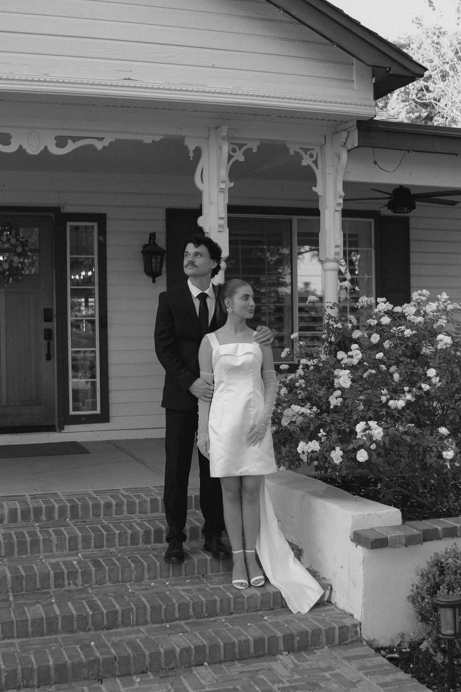 Black and white photo of a couple in wedding attire posing on brick steps outside a charming house with roses.