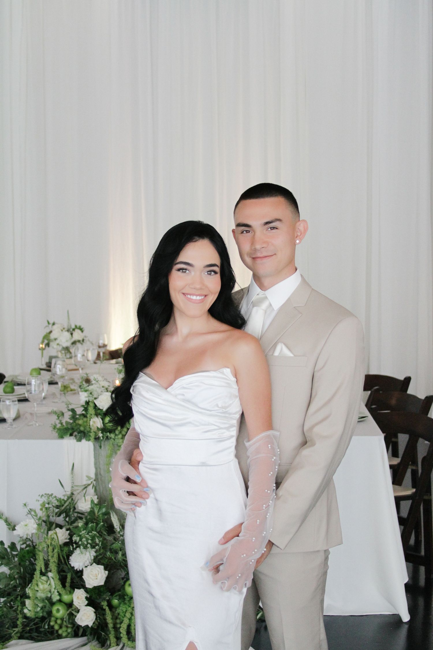 Bride in white strapless gown and groom in beige suit pose together at elegant wedding reception.
