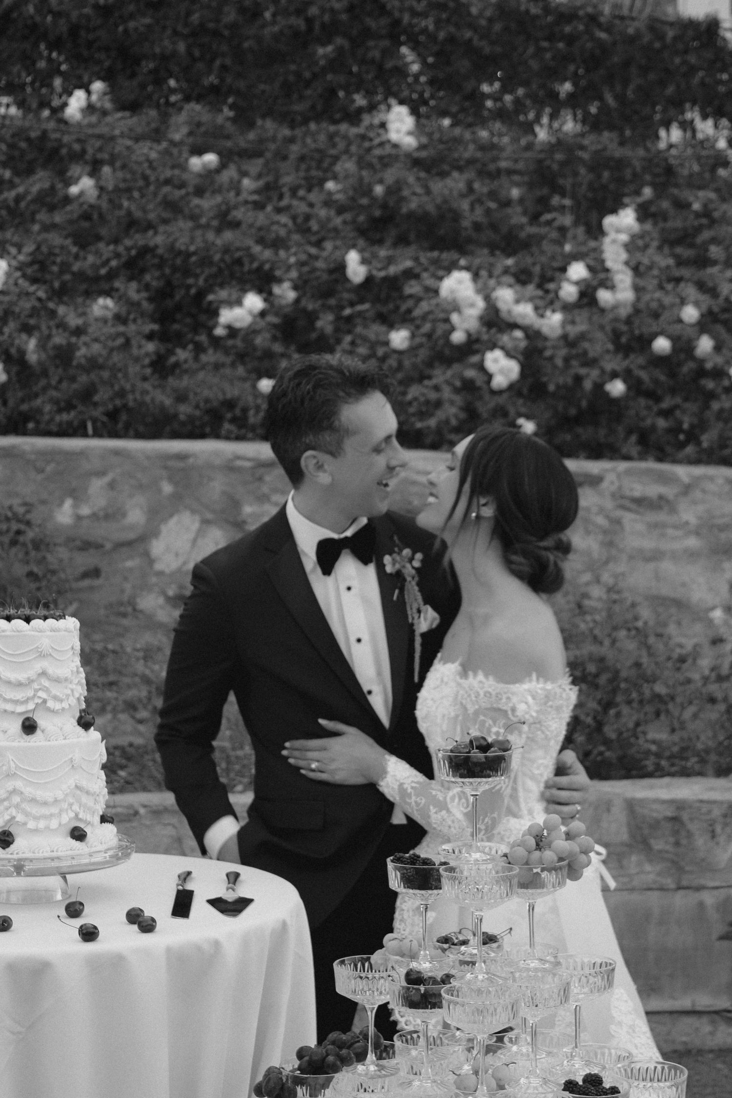 Black and white photo of bride and groom sharing a kiss beside wedding cake and champagne tower display.