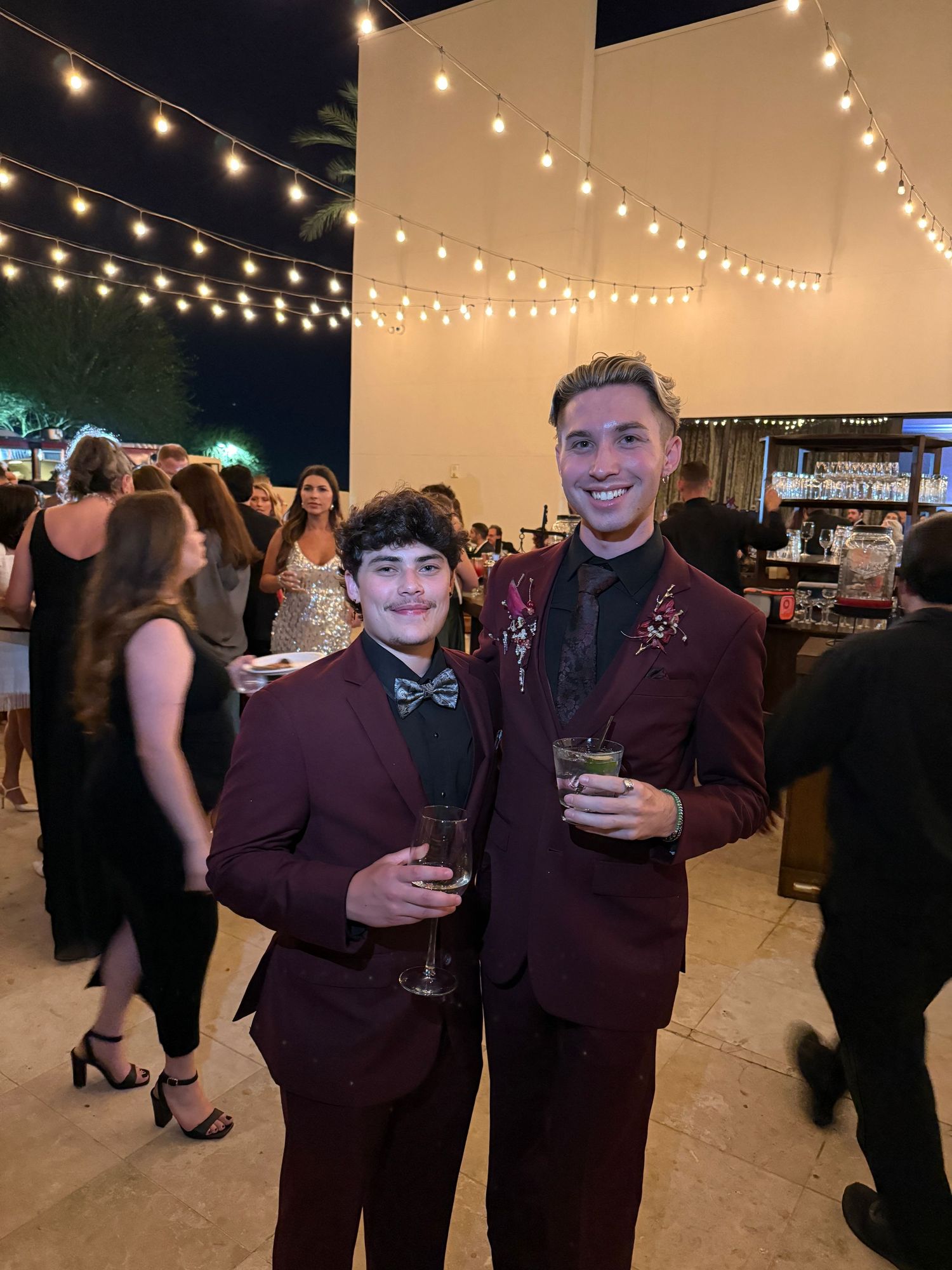 Two men in matching burgundy tuxedos holding drinks at an elegant outdoor evening wedding reception with string lights.