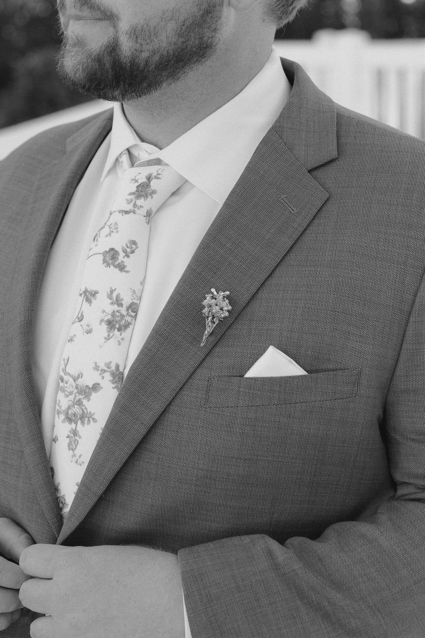 Close-up details of a grey suit jacket with white floral tie and pocket square in black and white photography.