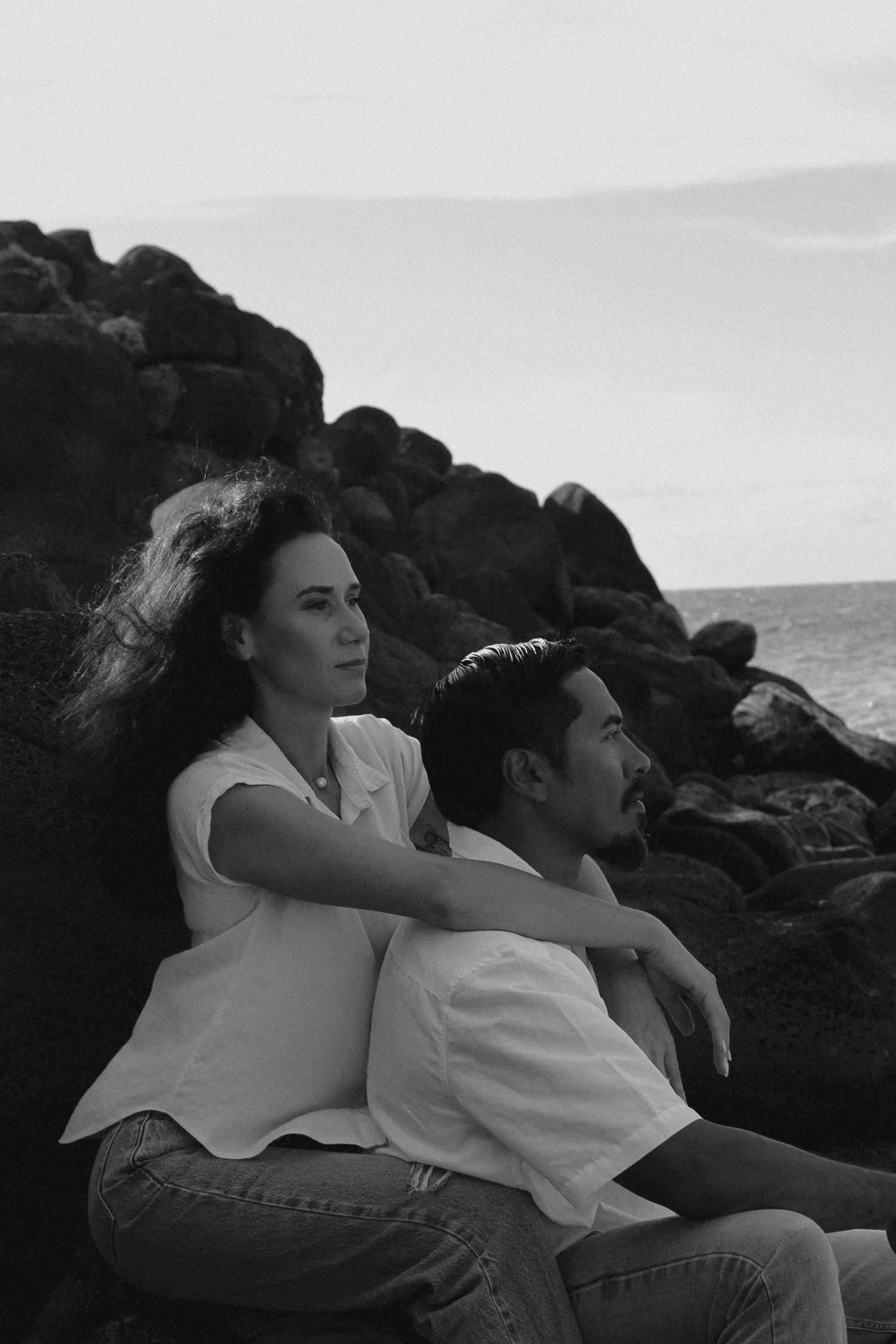 A couple sits on rocky coastal cliffs in black and white, enjoying a peaceful moment by the ocean together.