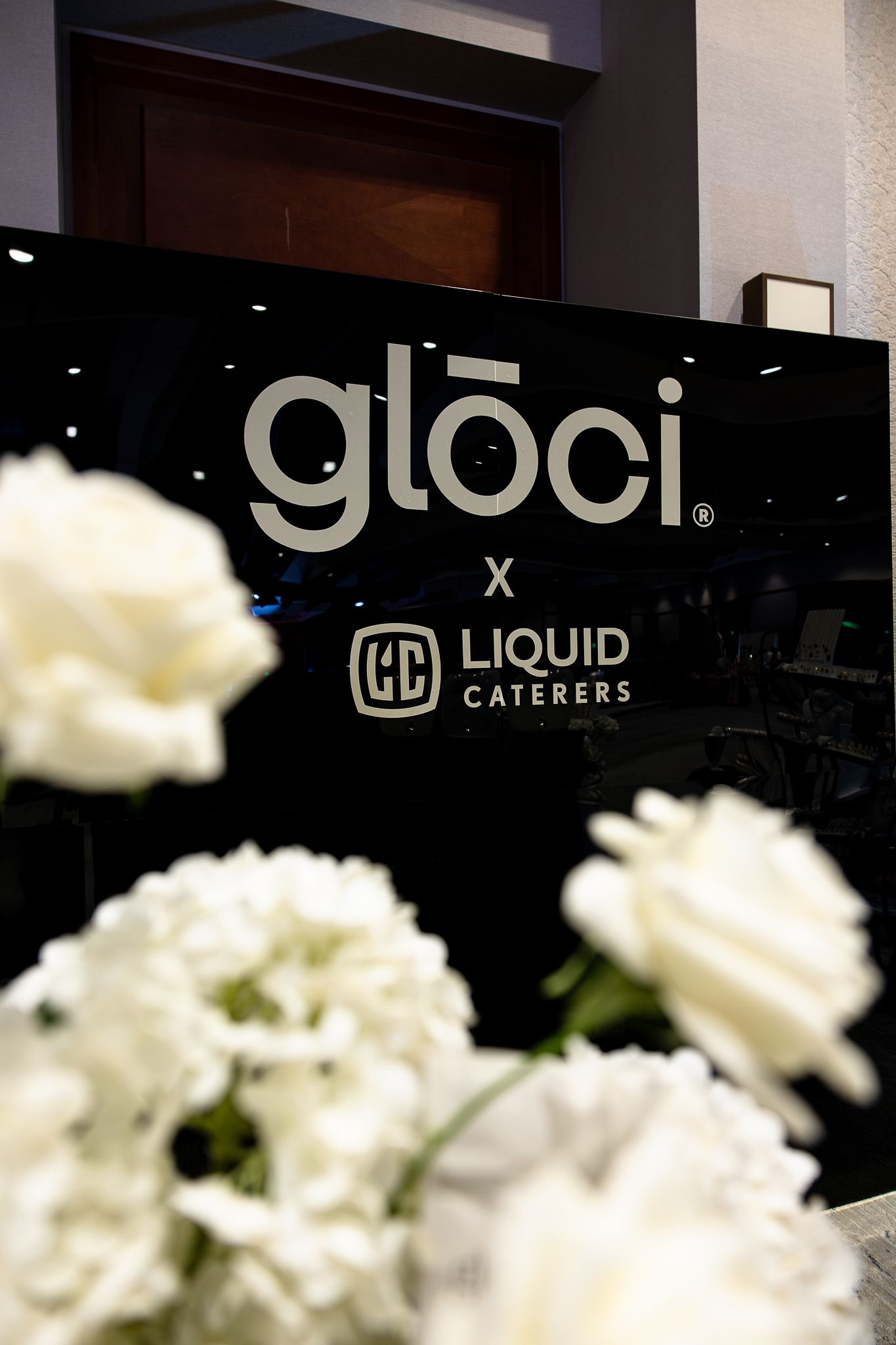Gloci x Liquid Caterers branded black sign displayed with white flowers at a catering event.