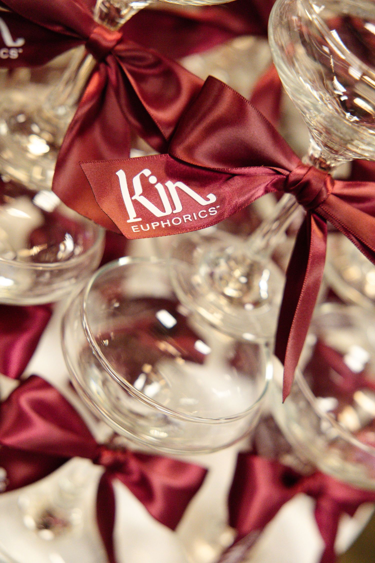 Crystal wine glasses adorned with burgundy satin bows and Kin Euphorics branded ribbon tags arranged elegantly.