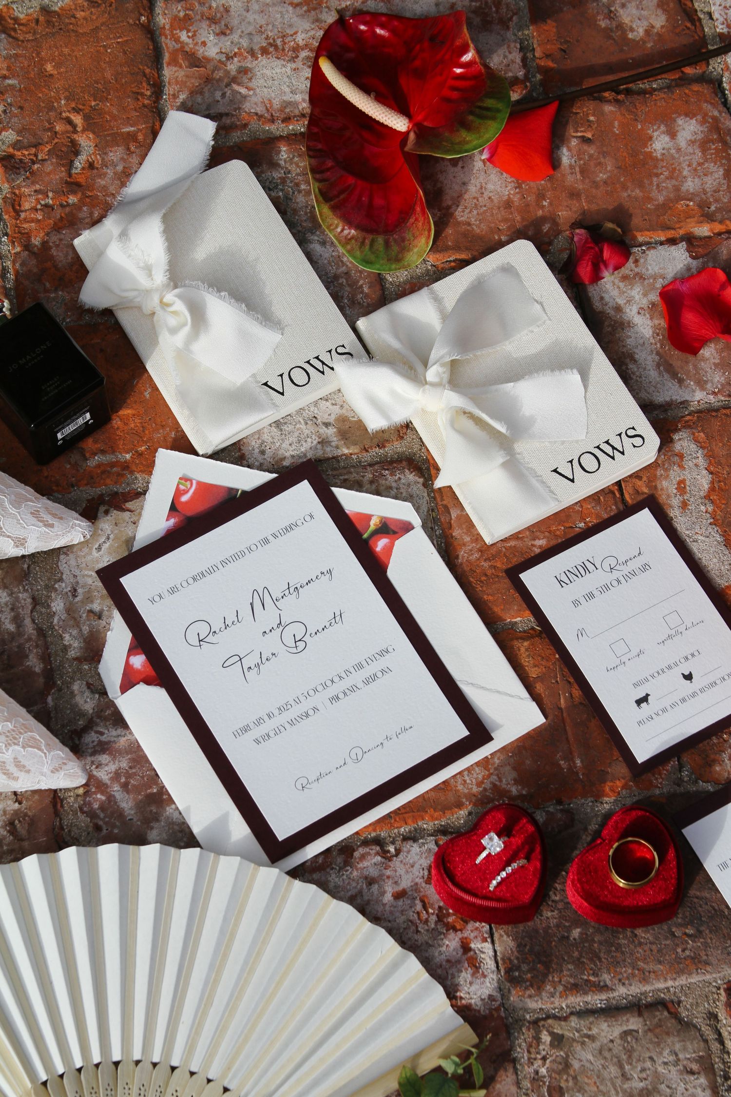 Wedding flat lay featuring invitation suite, vow books, red ring boxes, and white fan on brick surface.