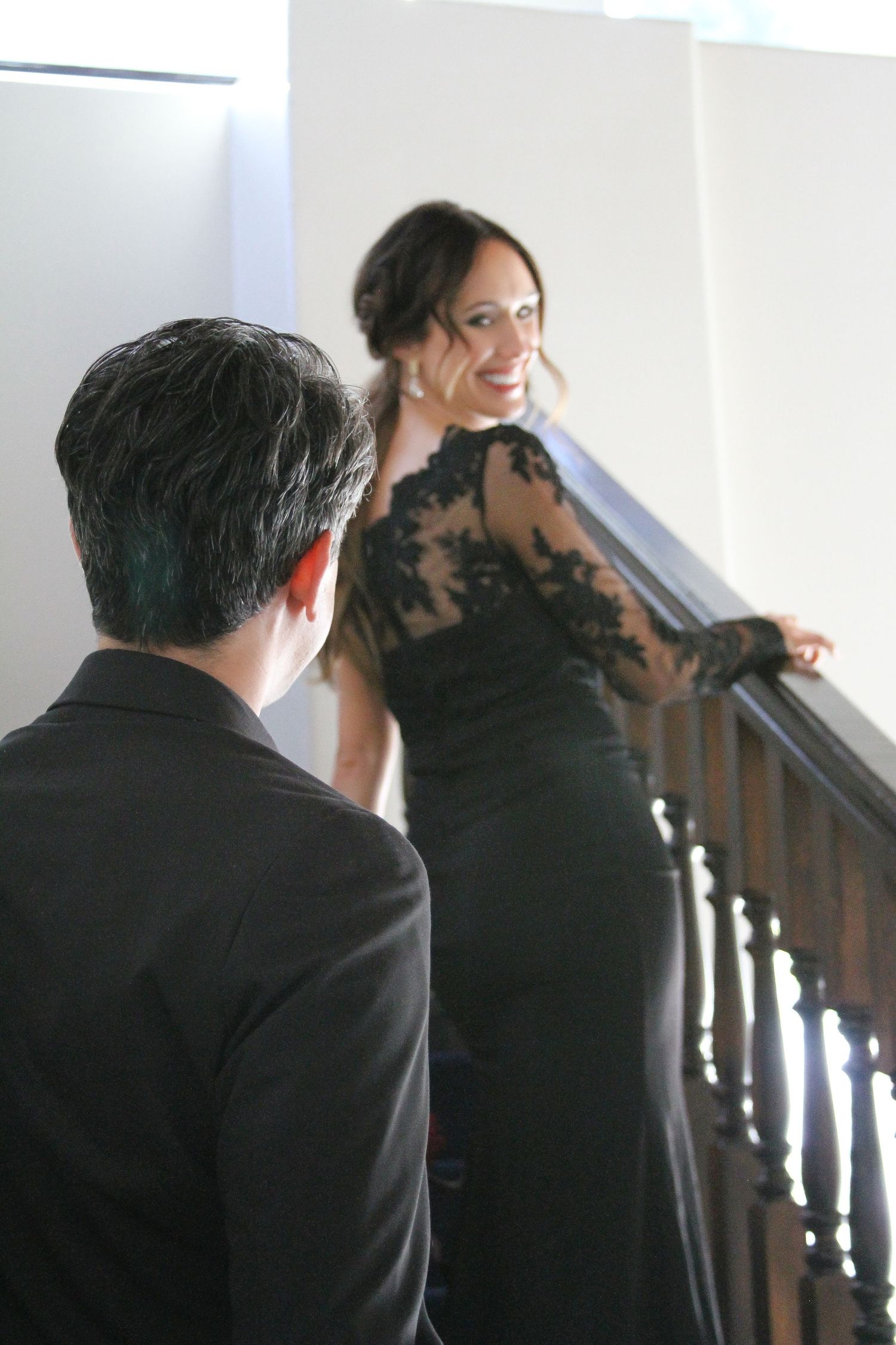 A woman in an elegant black lace evening gown stands on stairs while someone in a dark suit looks on.
