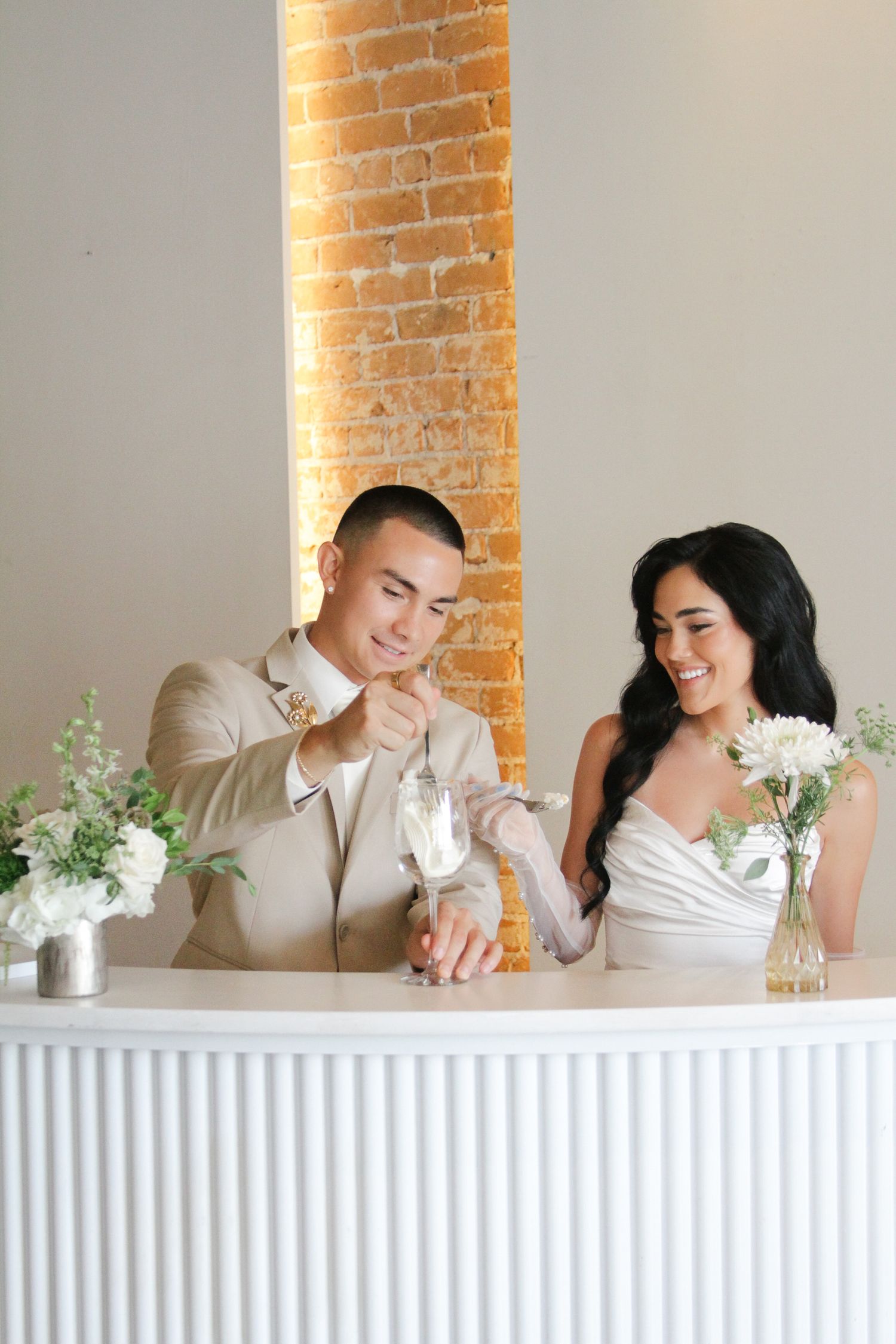 Smiling bride and groom in elegant attire at white bar with white floral arrangements.