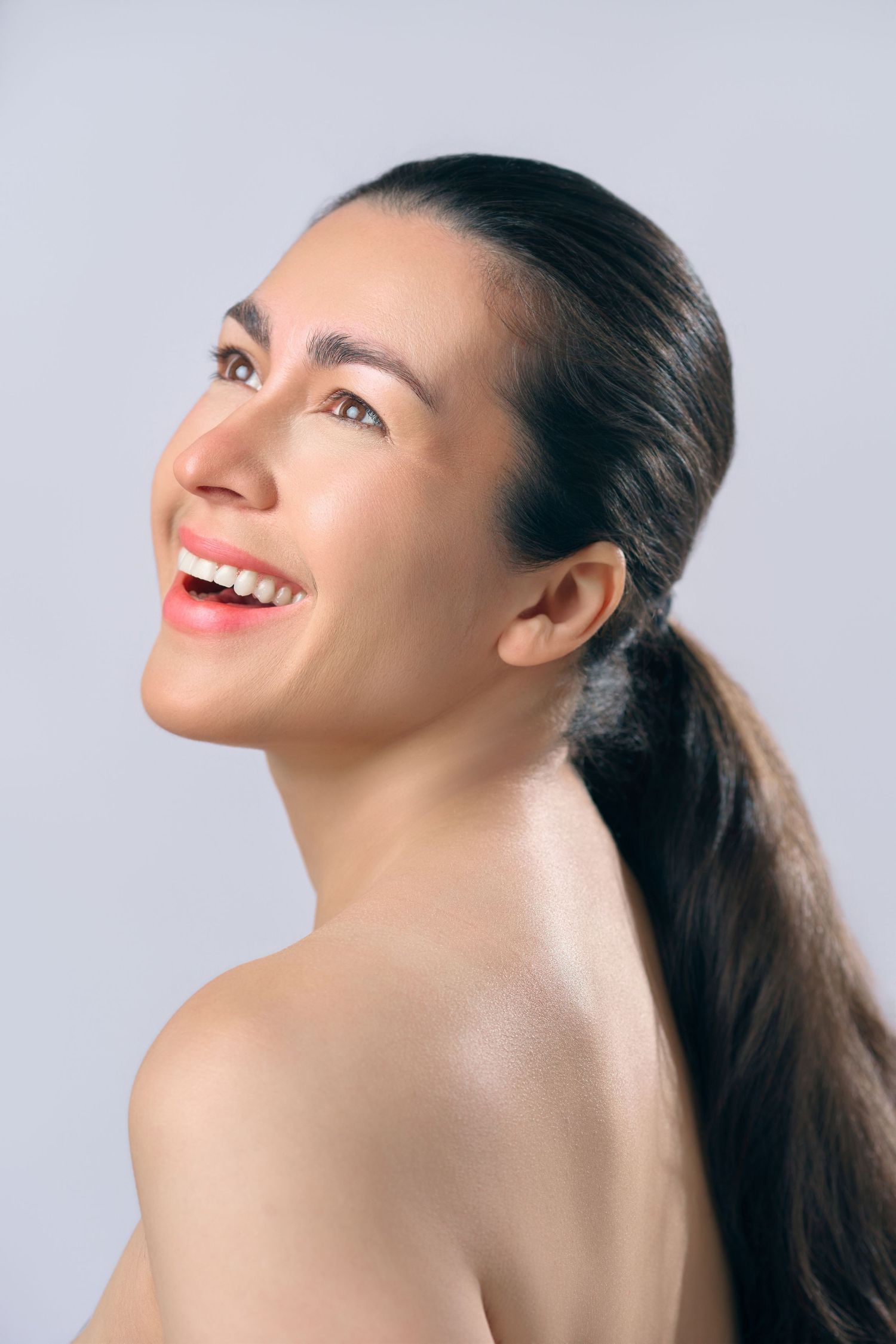 A beautiful smiling woman with dark hair in a ponytail looks up joyfully against a light gray background.