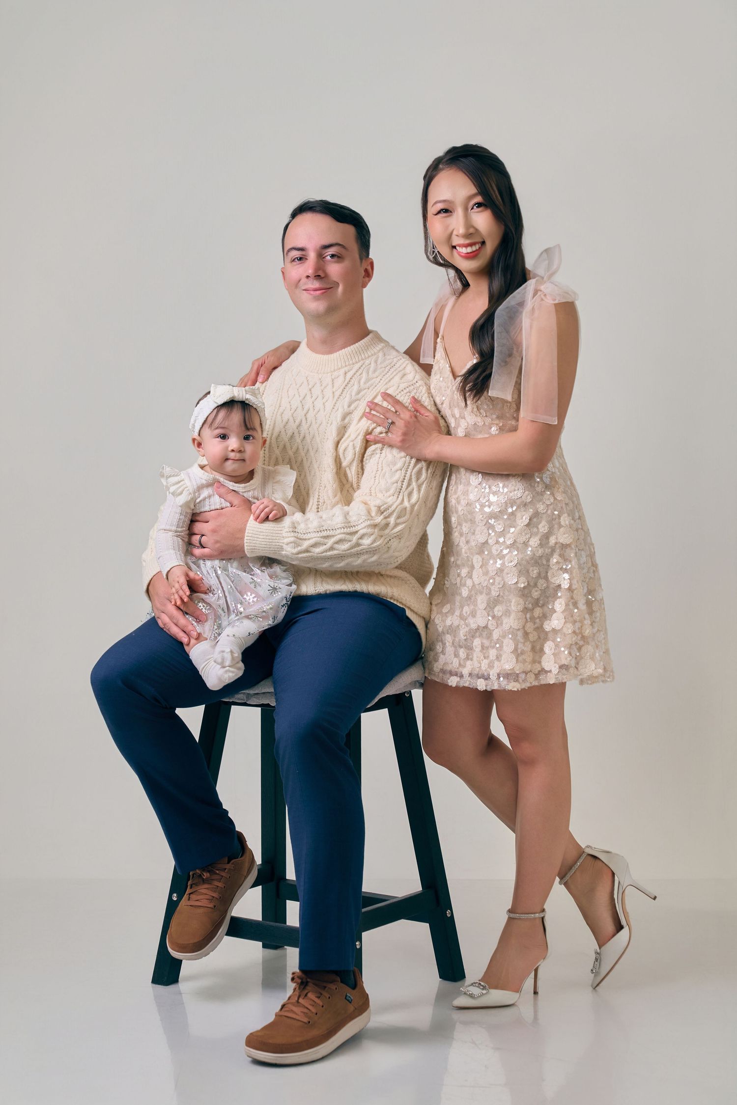 Young family studio portrait with father seated holding baby girl, mother in sequined dress standing beside them.