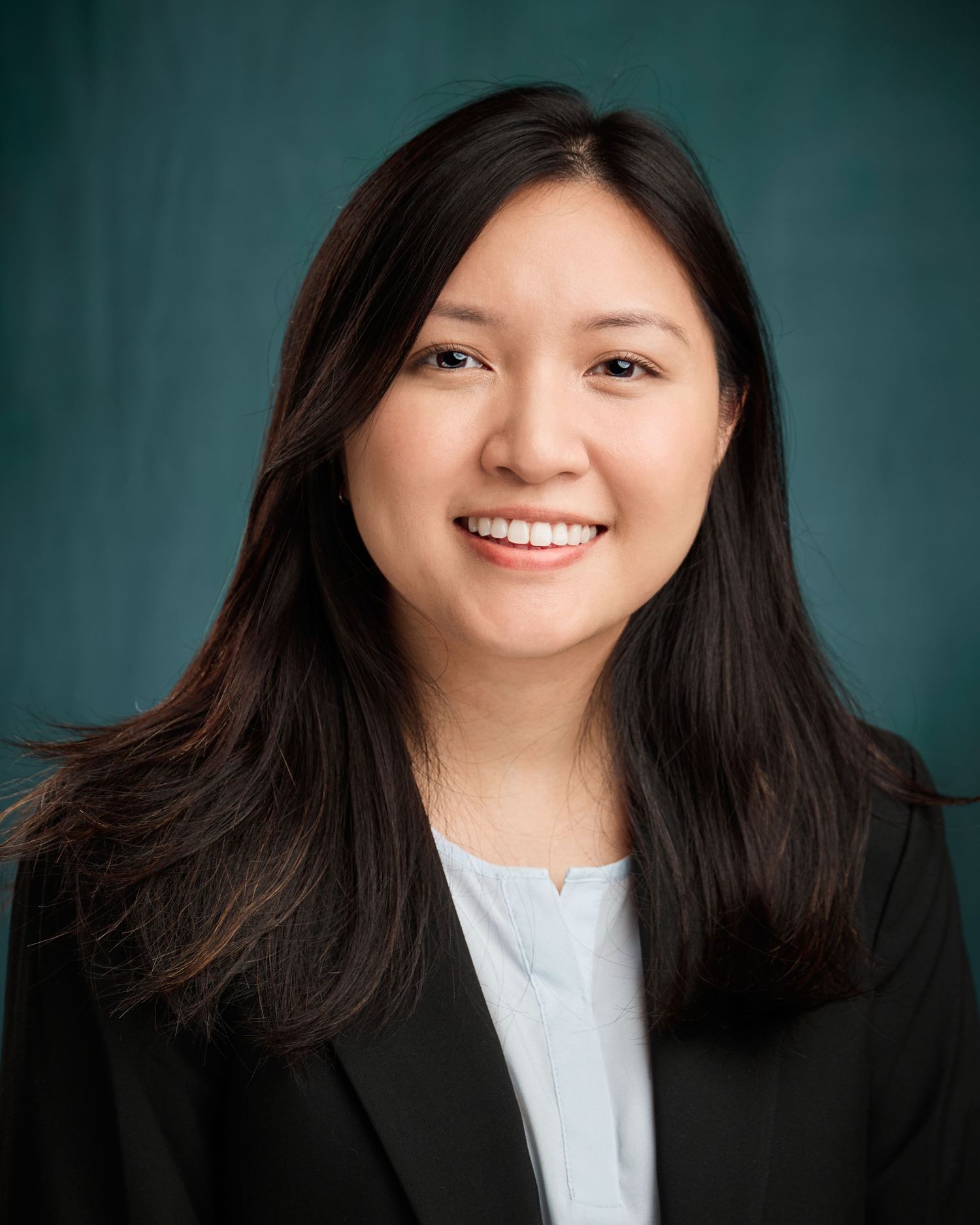 Professional headshot of a smiling Asian woman in a black blazer against a teal background.