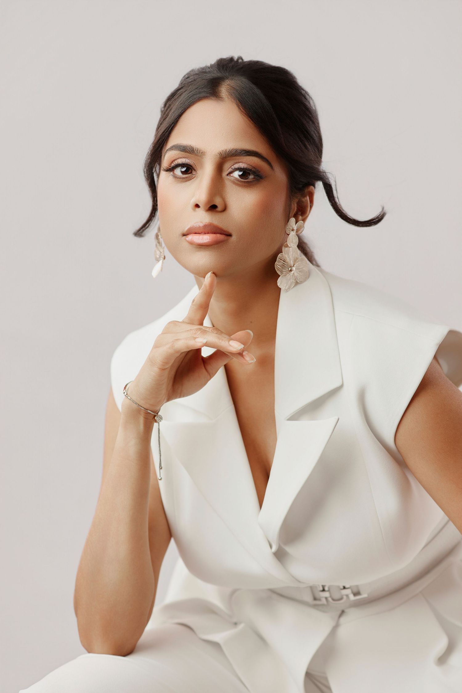 Elegant woman in a white blazer dress posing thoughtfully with hand on chin against a light background.