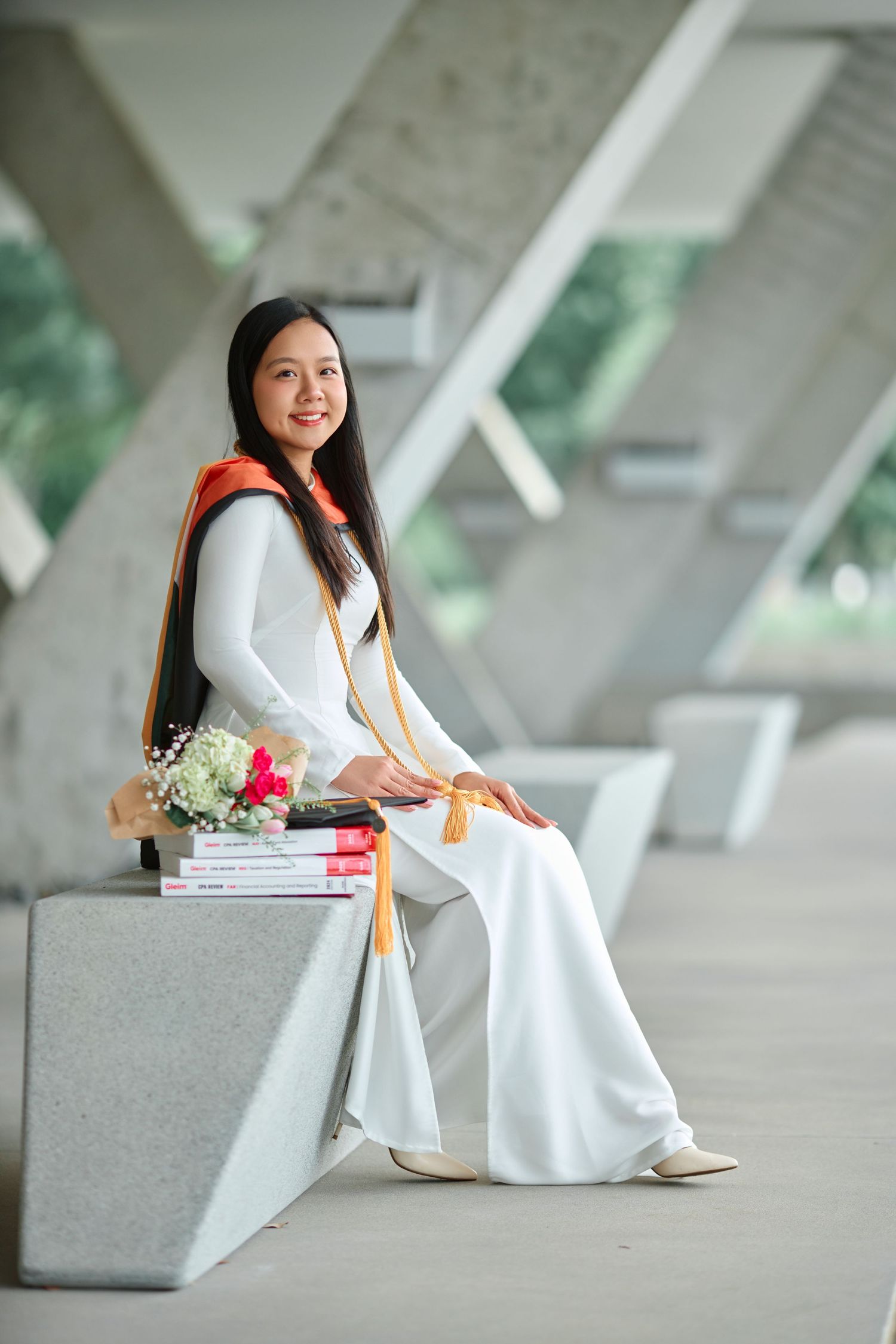 Vietnamese graduate in traditional white ao dai with orange stole sits on concrete bench.