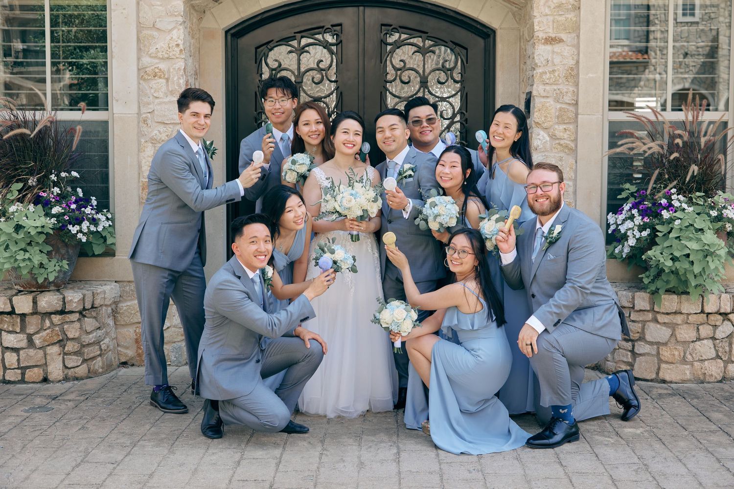 Wedding party in gray and blue attire celebrating with the bride and groom outside stone venue doors.