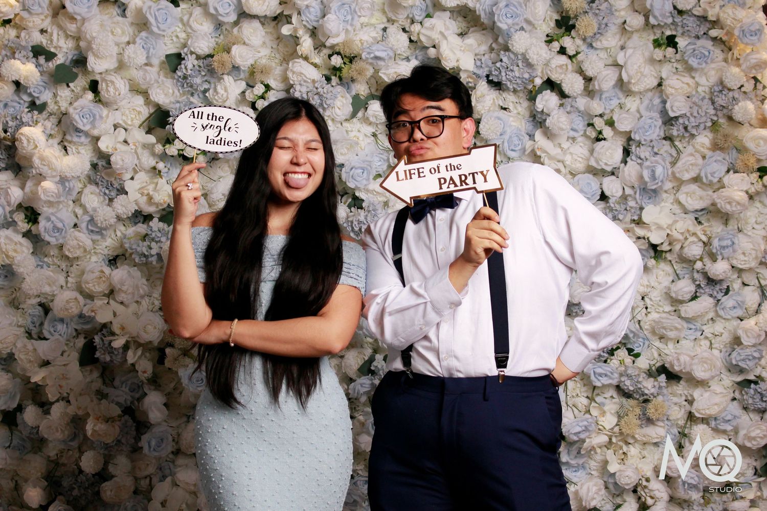 Two people posing at a floral photo booth with fun props, one holding a Life of the Party sign, against a white flower wall.