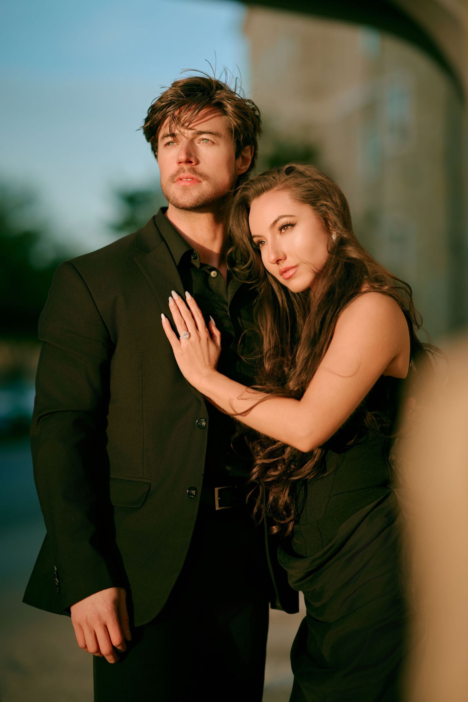 Elegant couple in black outfits posing outdoors, woman with long hair leaning against man in suit at dusk.