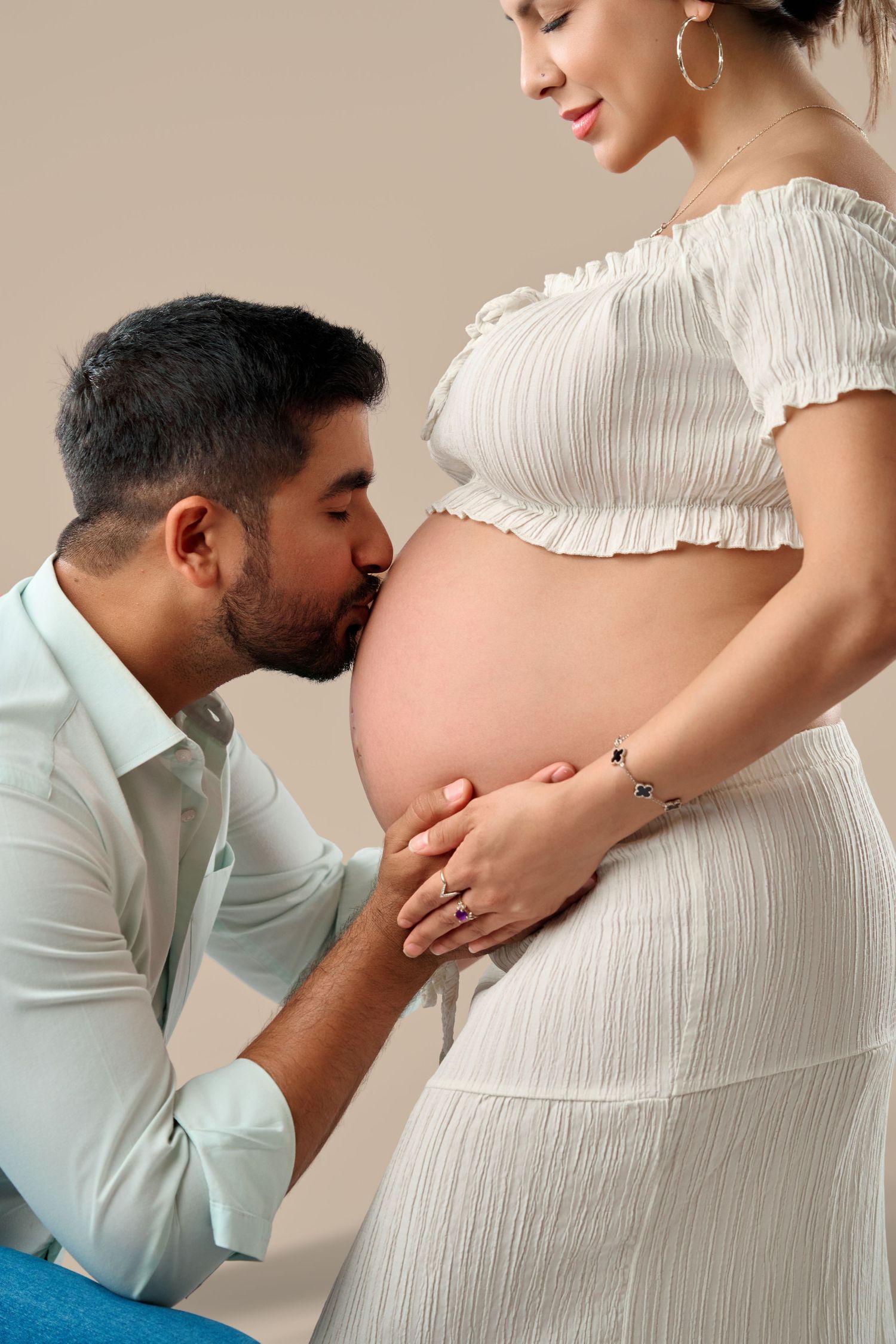 A man kisses his pregnant partner's bare belly while she smiles, both dressed in white during a maternity photoshoot.