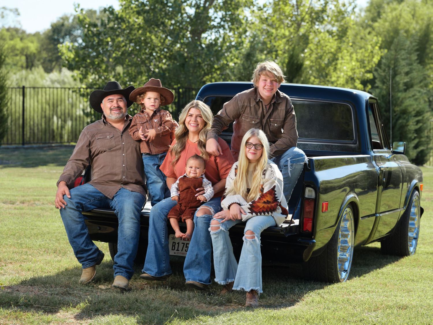A happy family of six posing together on the back of a vintage black pickup truck in a green field.